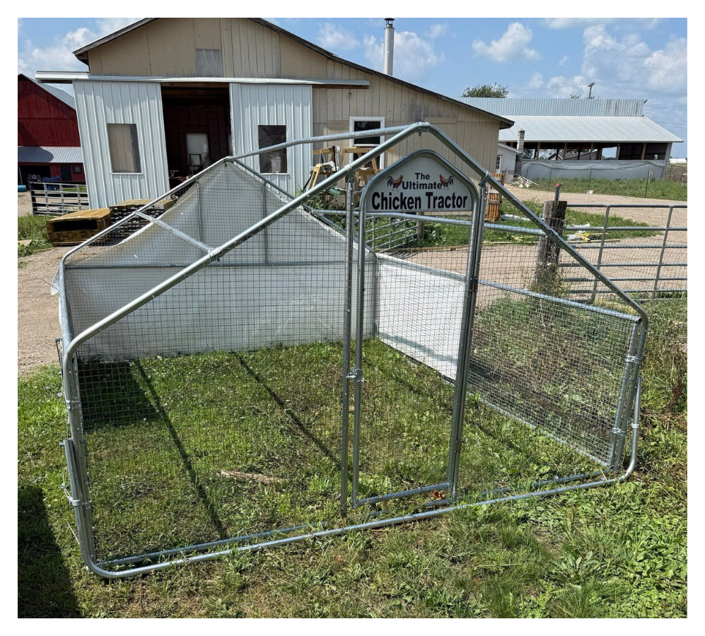 A small metal chicken coop with a sign that says 'The Ultimate Chicken Tractor.' It is located outdoors on grass near farm buildings.