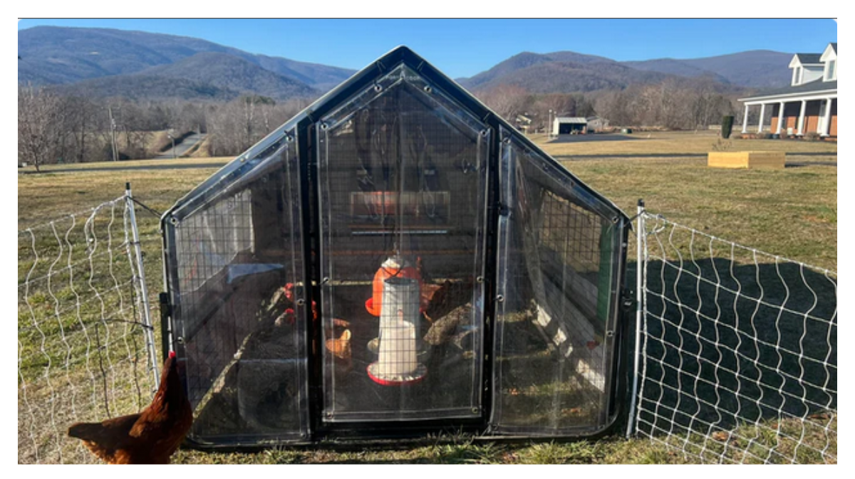 A small chicken coop with a wire fence on a grassy field, mountains in the background, and a house on the right side.
