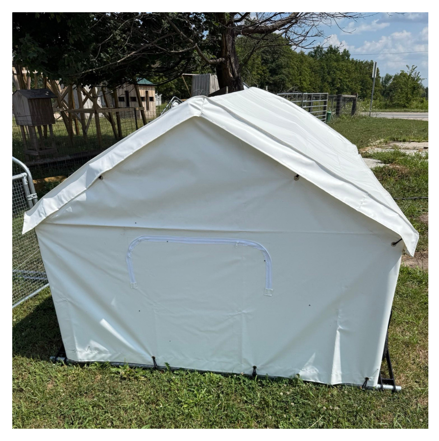 A white outdoor tent with a zippered opening, set up on grass, with trees, a wooden fence, and a farm or zoo background.