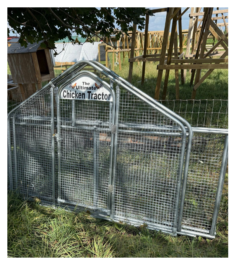 A metal chicken tractor with a sign reading 'The Ultimate Chicken Tractor,' situated outdoors in a grassy area with construction or farm structures in the background.