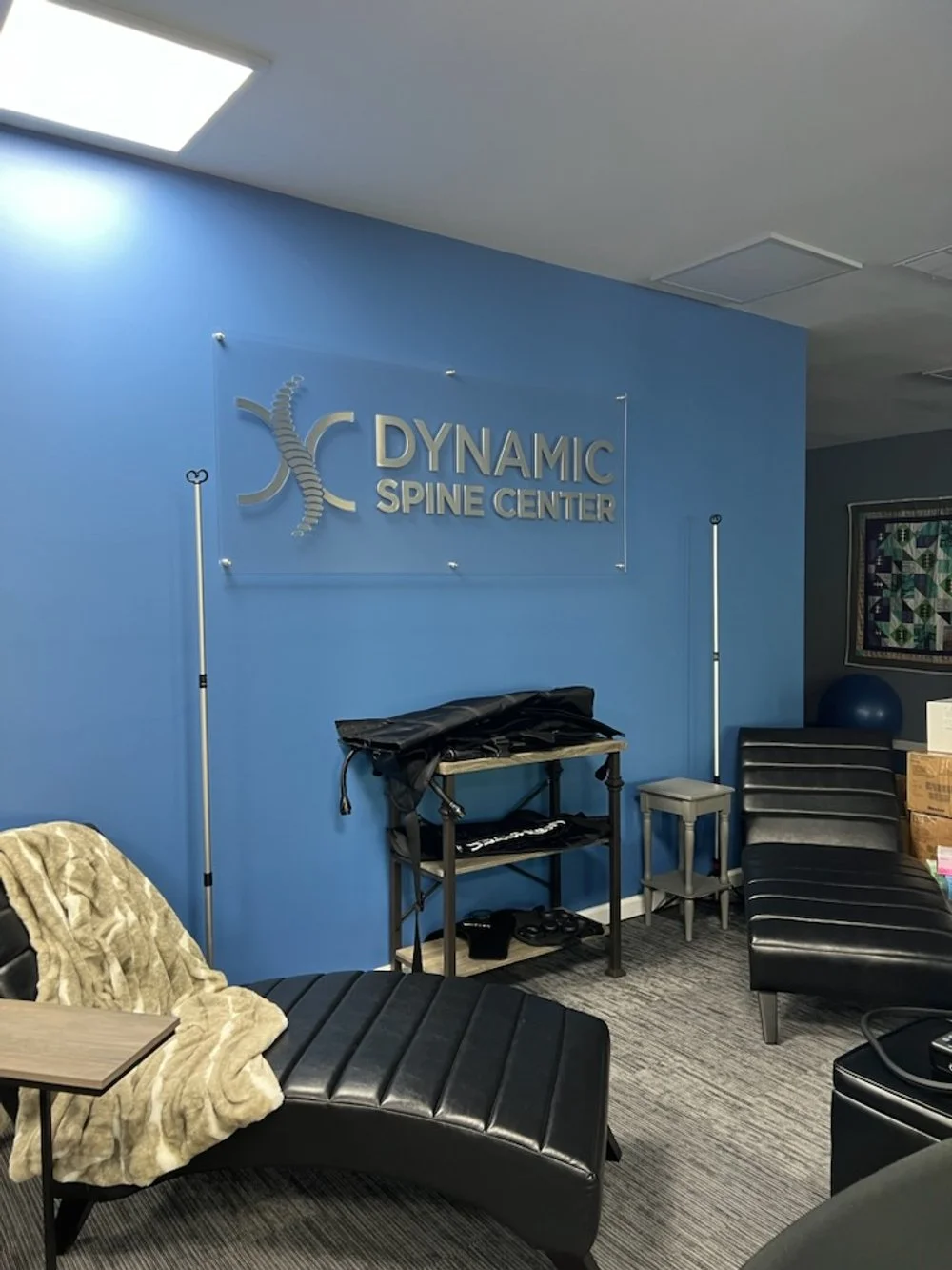 Interior of a spine center with blue walls, black lounge chairs, a small table, and a blanket. A sign reads 'Dynamic Spine Center.'