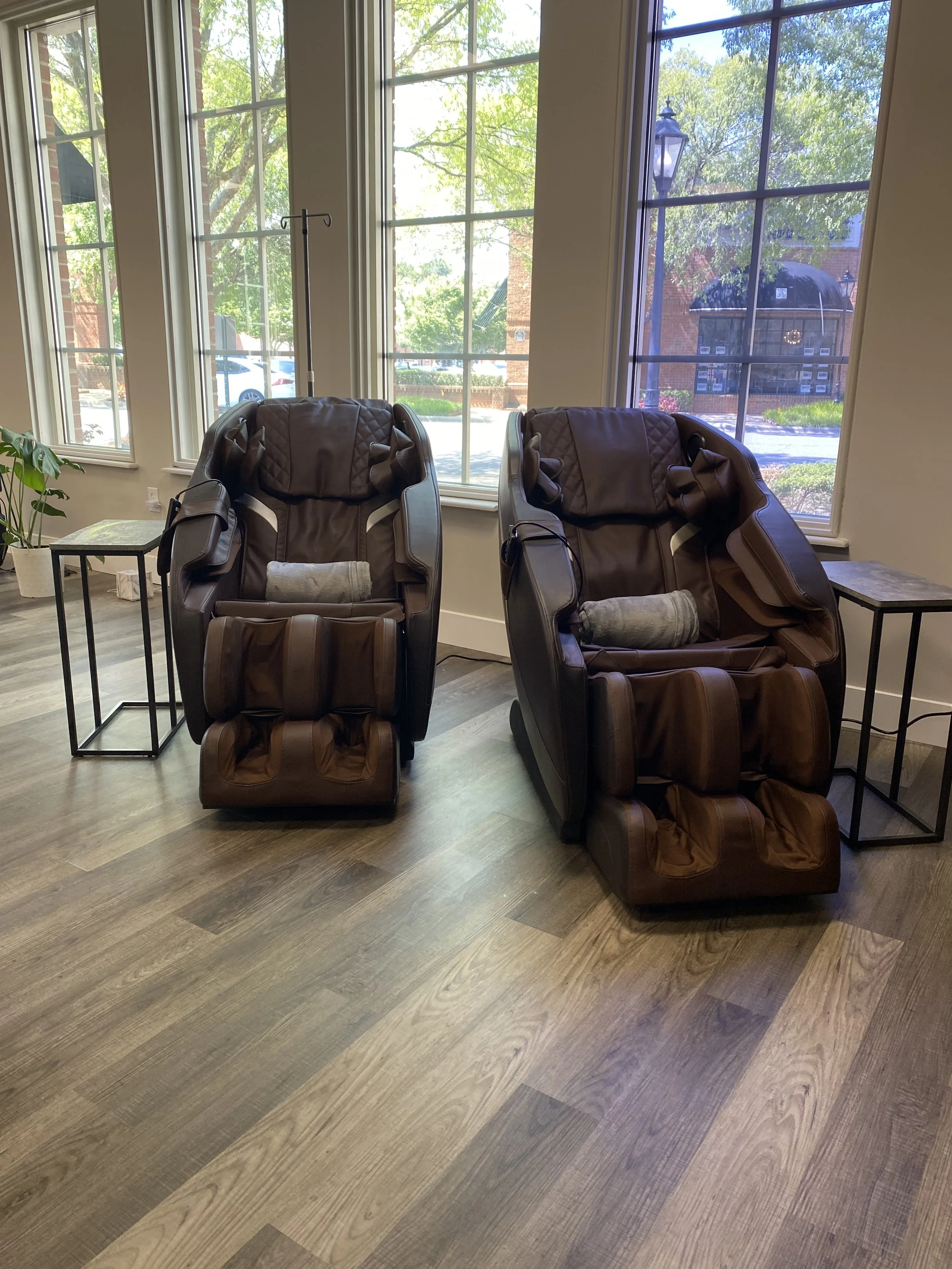 Two brown massage chairs positioned near large windows in a room with wooden flooring.