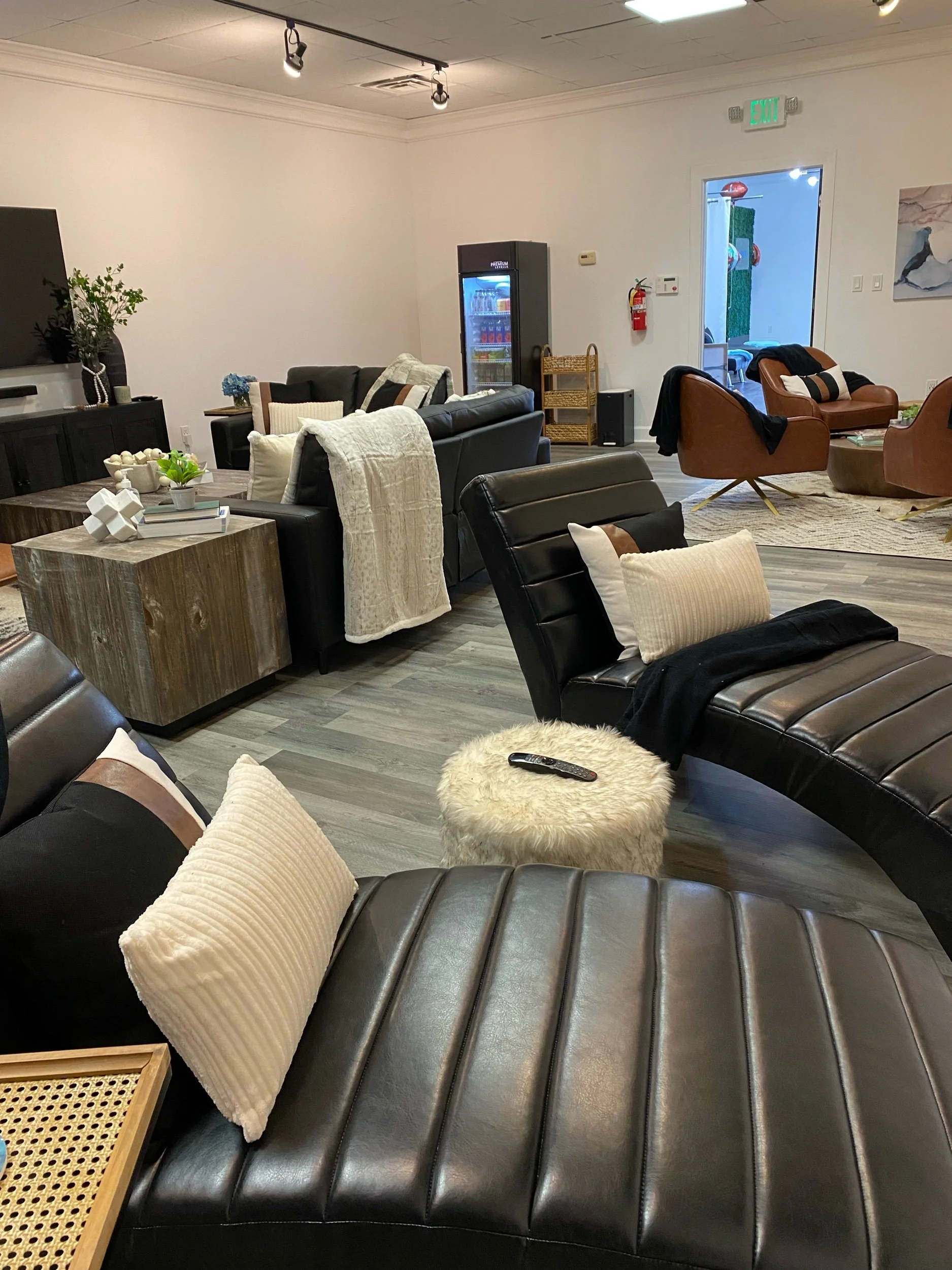 Modern lounge area with black leather chairs, a beige ottoman, various decorative pillows, a wooden coffee table, a mini fridge, and plants on a wooden floor.