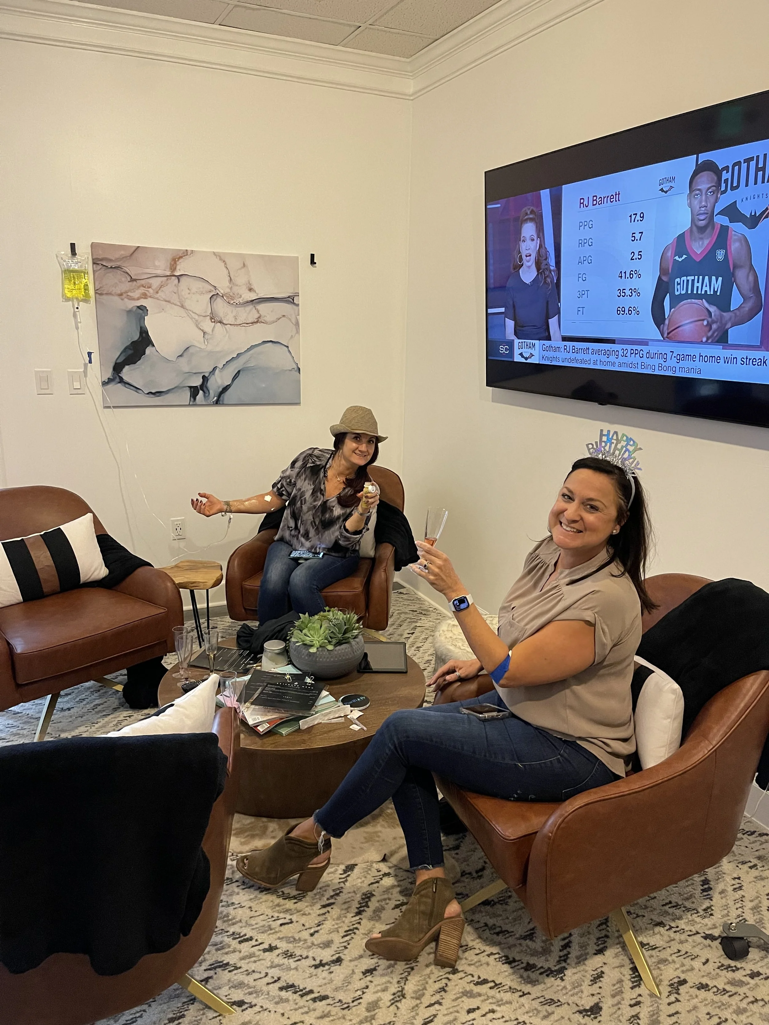 Two women sitting in leather chairs in a lounge area, one with an IV attached, holding drinks and smiling. A television on the wall shows a sports broadcast with player statistics. A round table with books and a plant is in the center.