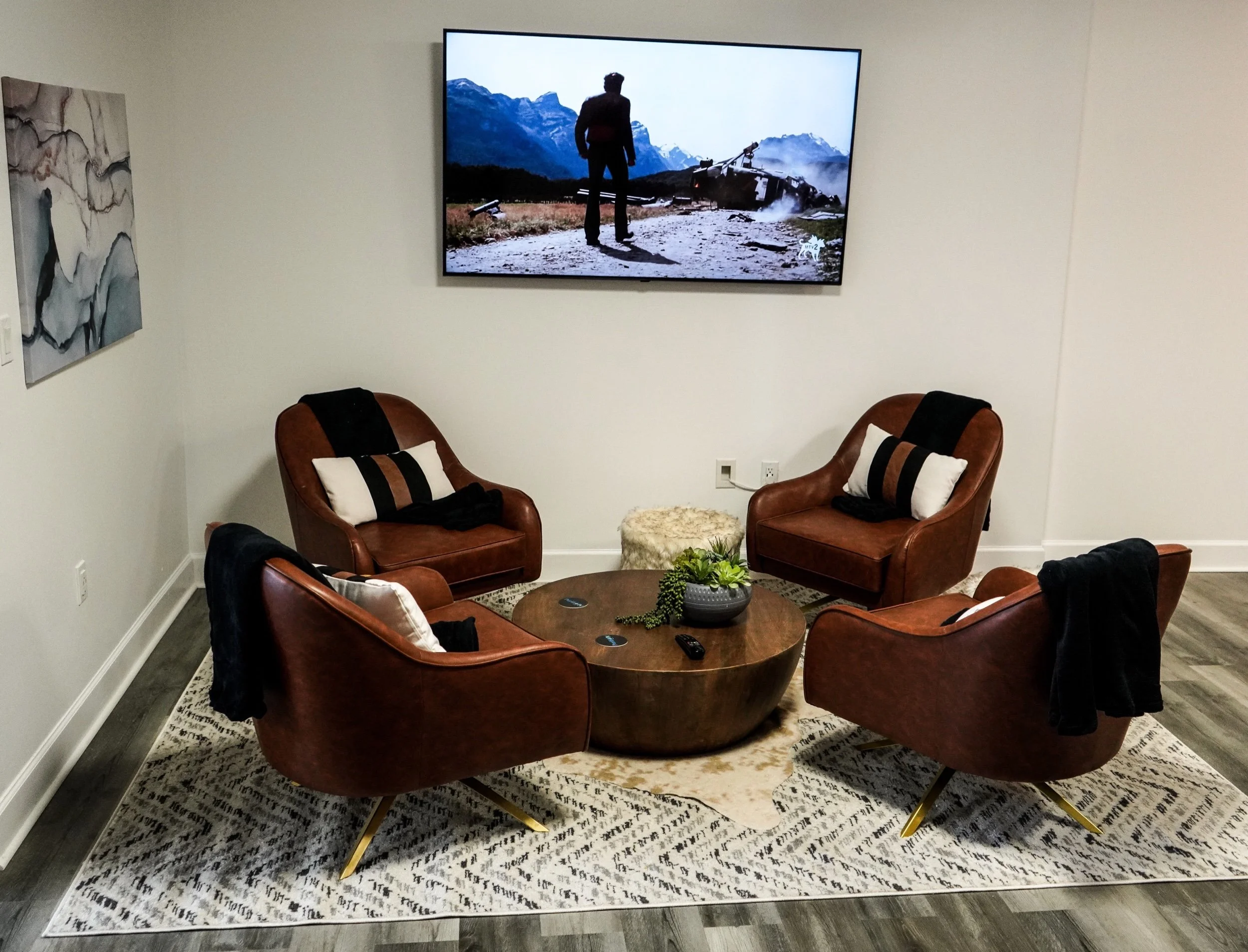 Stylish living room with four brown leather chairs arranged around a round wooden coffee table. A plant and remote control are on the table. Wall-mounted TV displaying a man silhouetted against mountain scenery. Modern decor includes abstract wall ar