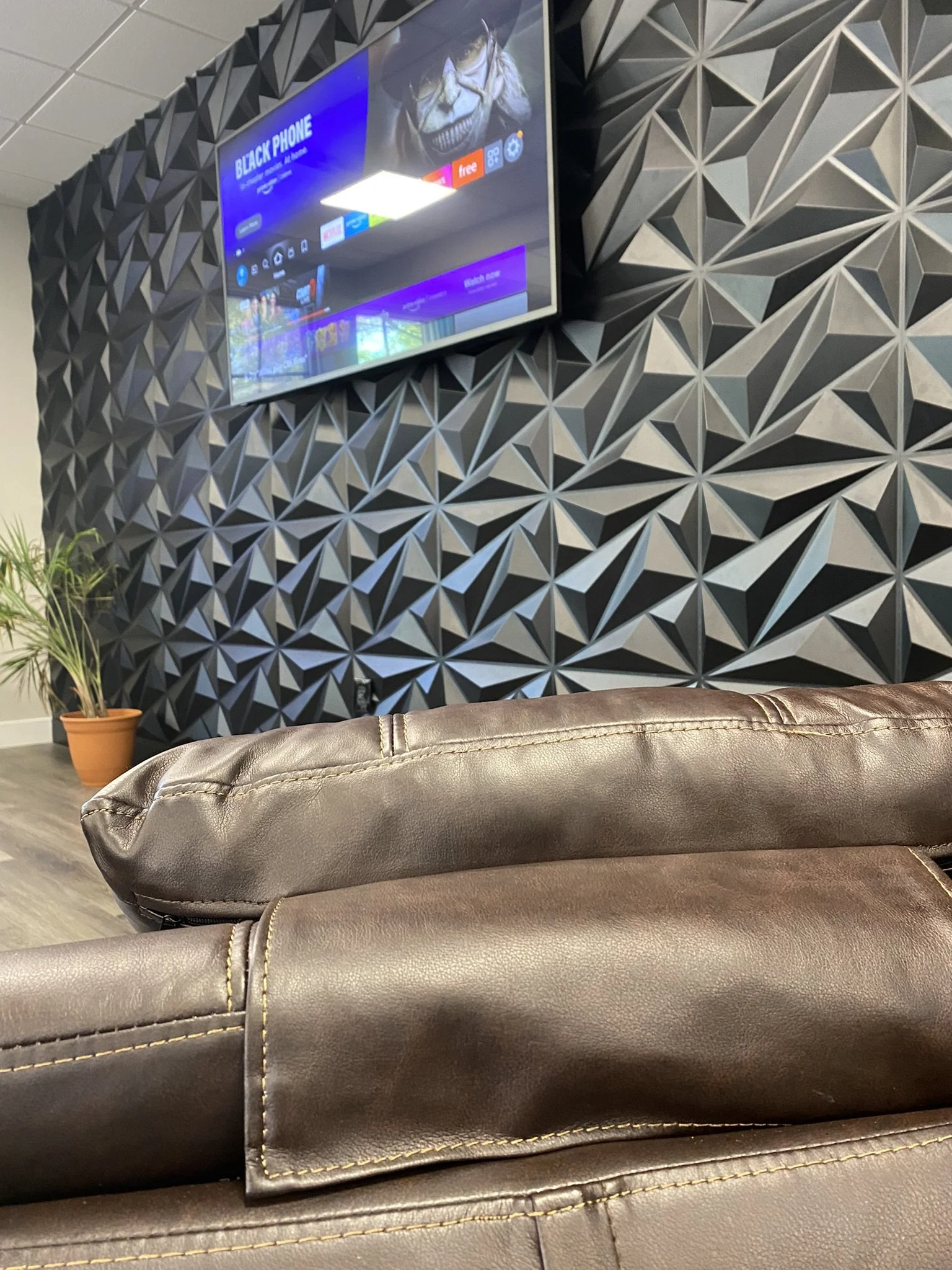 Leather couch in foreground, geometric patterned wall, mounted TV displaying "Black Phone" content screen, potted plant on wooden floor.