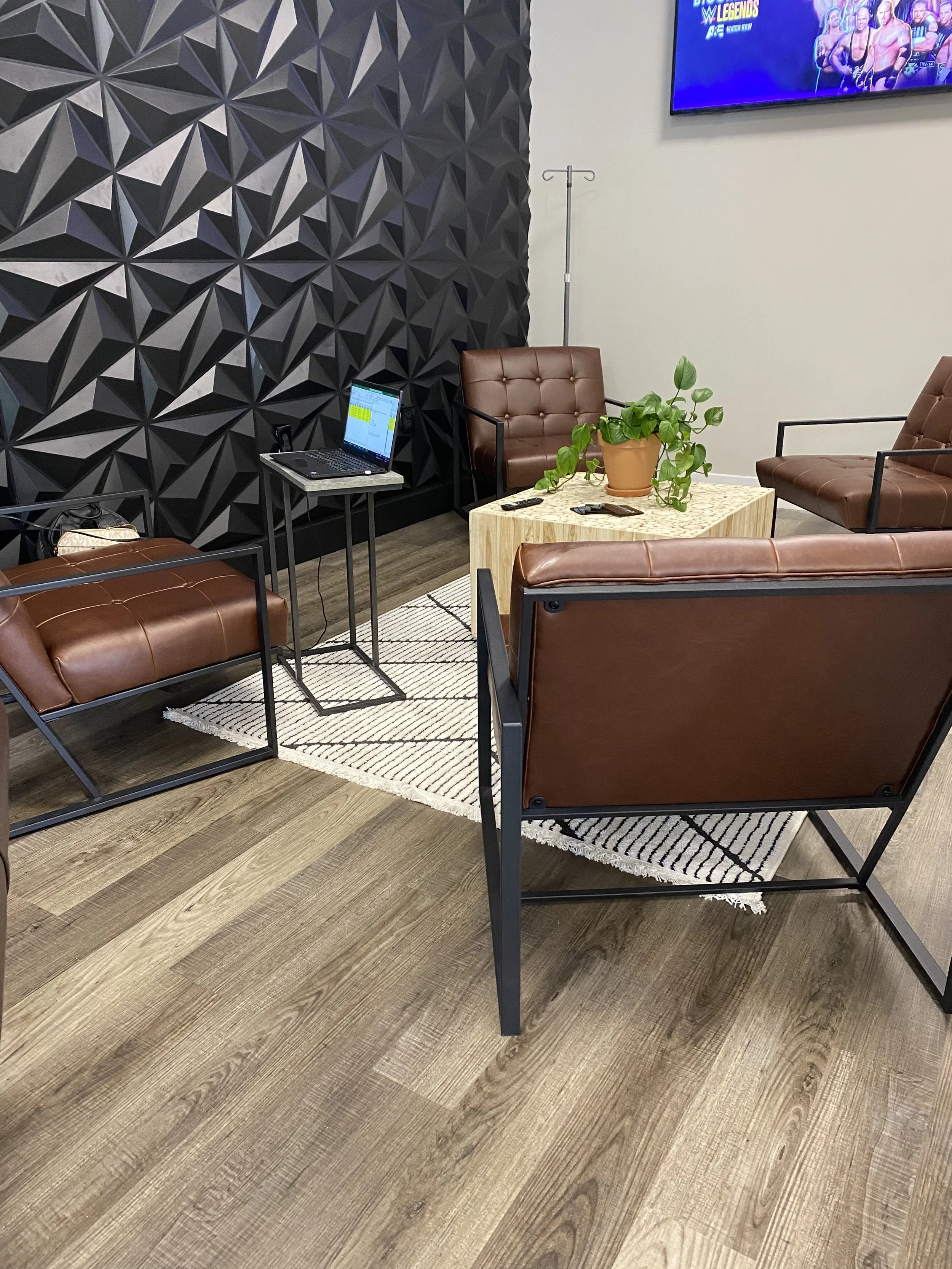 Modern office lounge with brown leather chairs, geometric black wall, small wooden table with a plant, laptop on stand, and TV with wrestling show on wall.