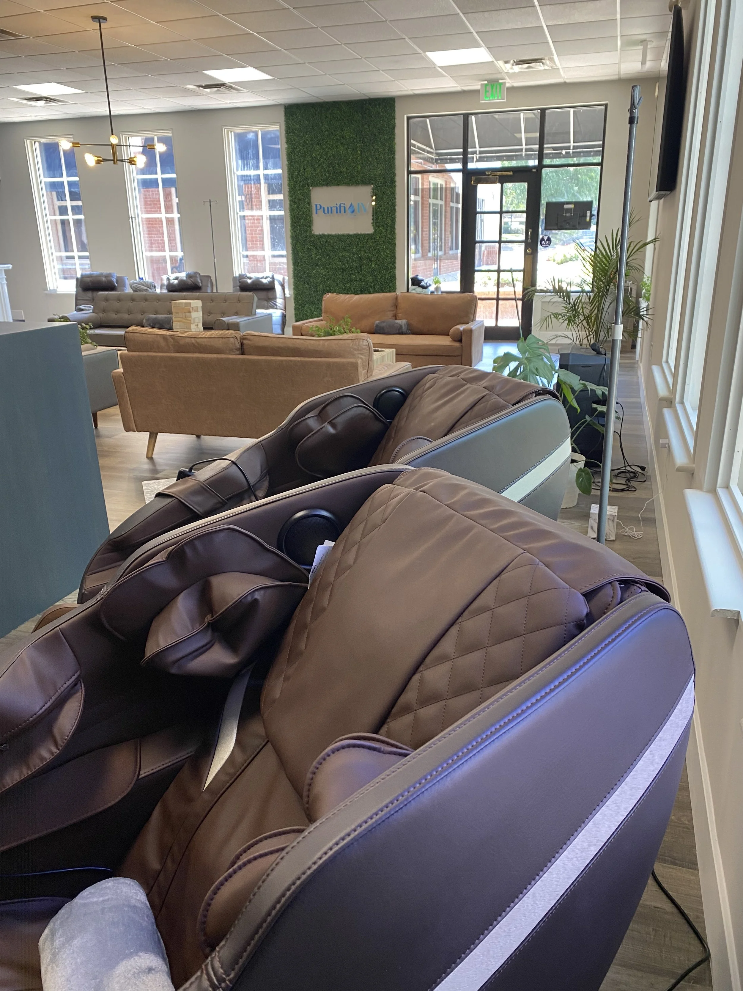 Interior of a modern lounge area with massage chairs, couches, a green wall, and large windows. The space appears well-lit and welcoming, with a sign saying "Purifit."