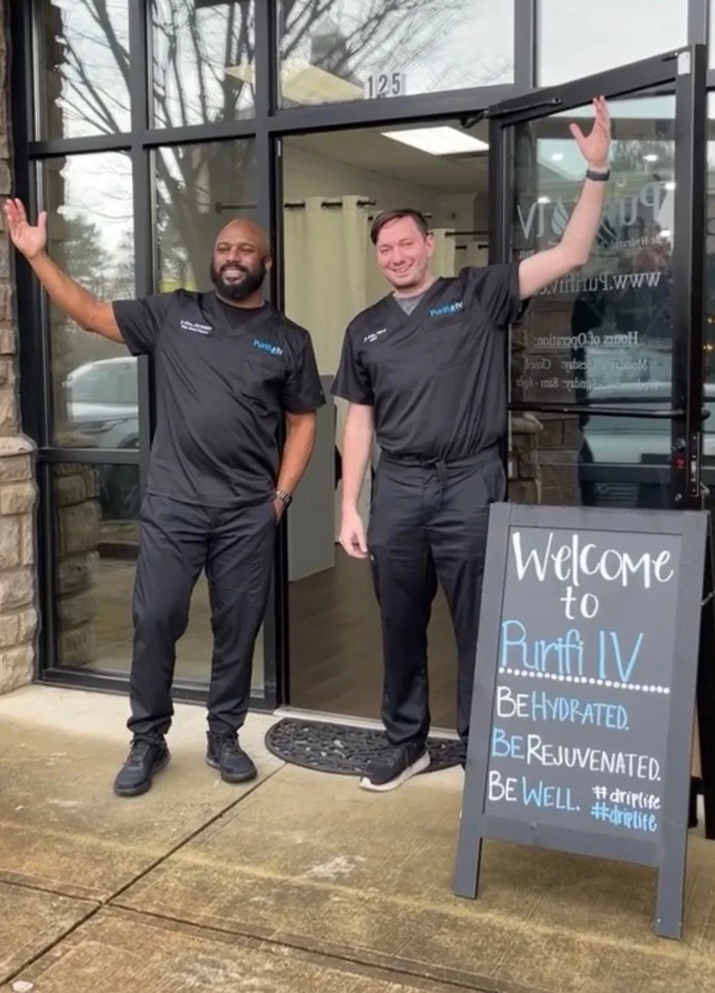 Two men in black uniforms standing outside Purifi IV clinic with a chalkboard sign that says "Welcome to Purifi IV. Be Hydrated. Be Rejuvenated. Be Well."