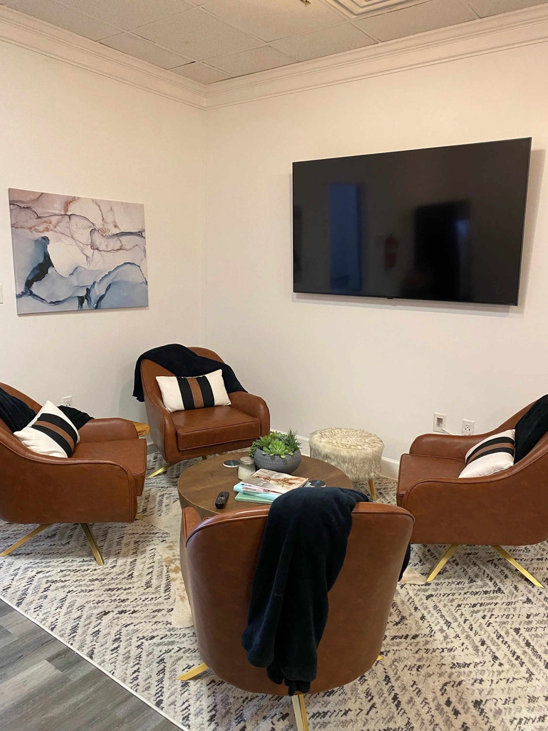 A modern living room with brown leather chairs, a round wooden coffee table, a flat-screen TV mounted on a white wall, abstract wall art, and a geometric-patterned rug.