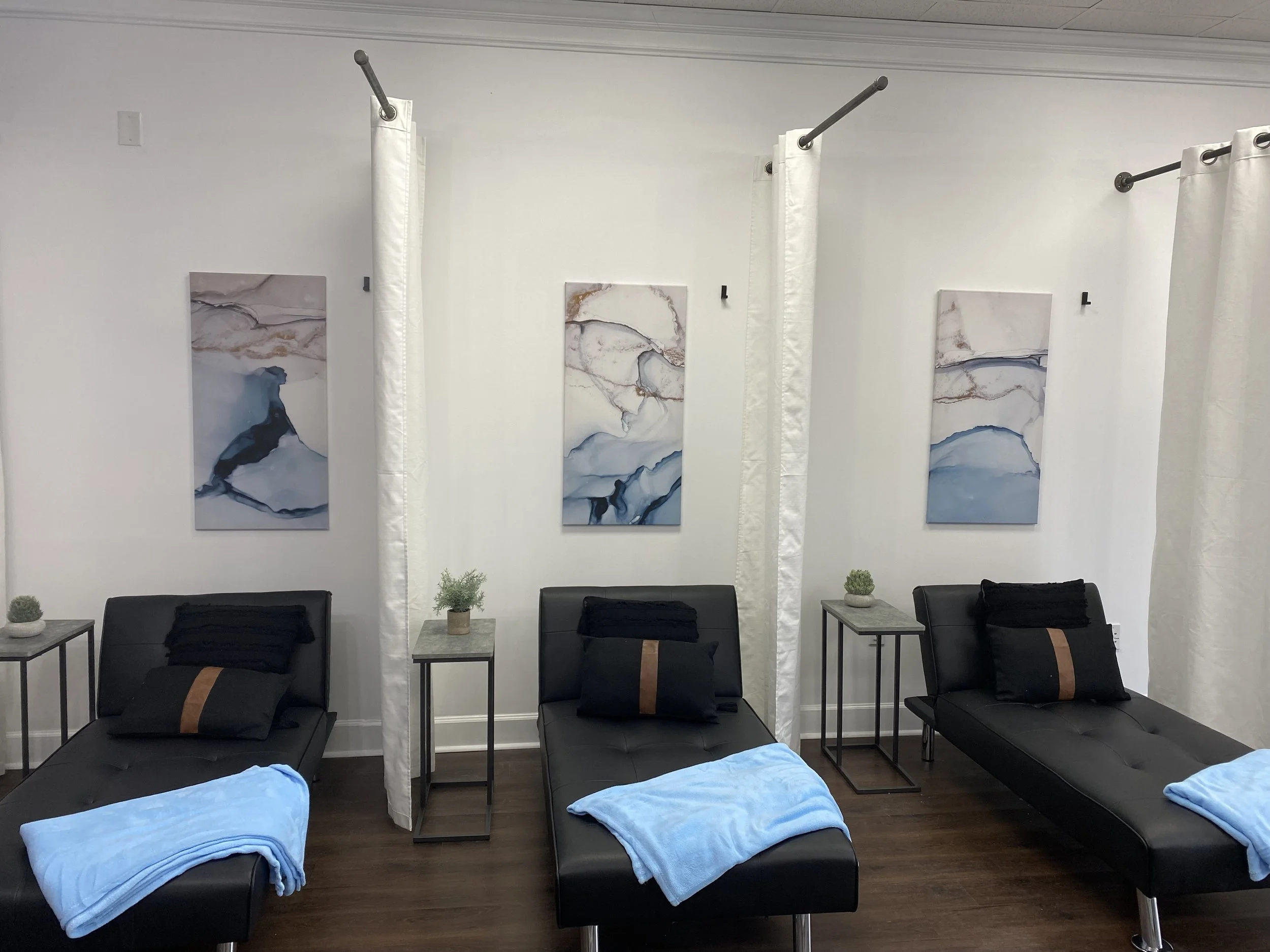 Interior of a spa with three black lounge chairs, each with a folded blue towel and black pillows. The room features white walls with abstract wall art and small side tables holding potted plants. White curtains partially separate the spaces.