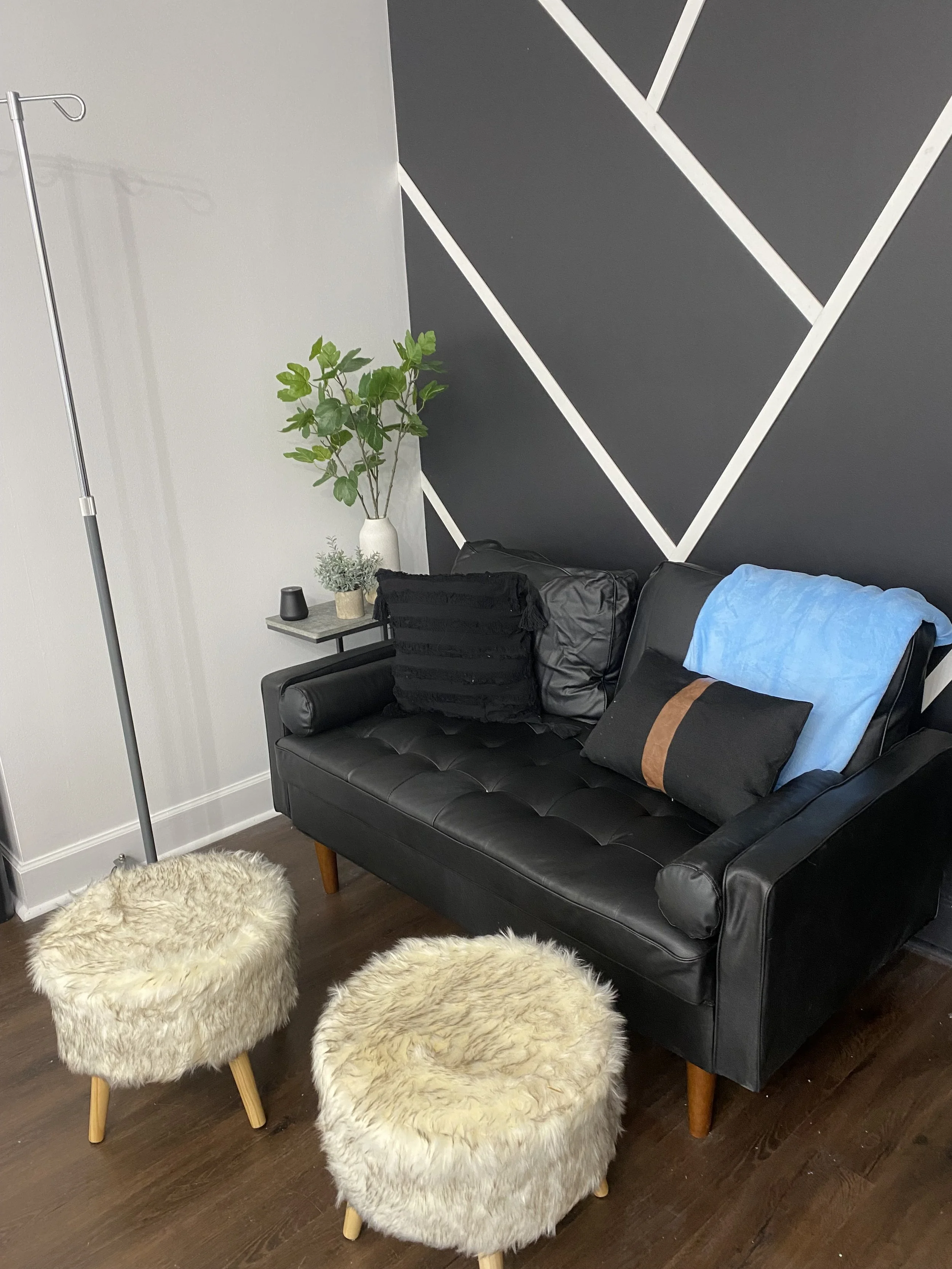 Modern living room corner with a black leather sofa, decorative pillows, two furry stools, a geometric black and white accent wall, a small side table with a plant, and an IV stand in the background.