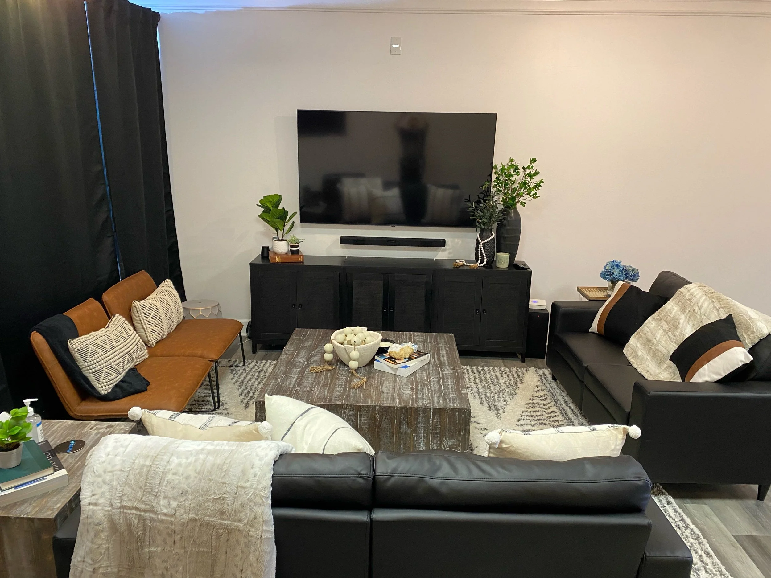 Modern living room with black couches, a brown loveseat, decorative pillows, a large flat-screen TV on the wall, a rustic wooden coffee table, houseplants, and various decor items. Black curtains and a gray area rug accent the space.