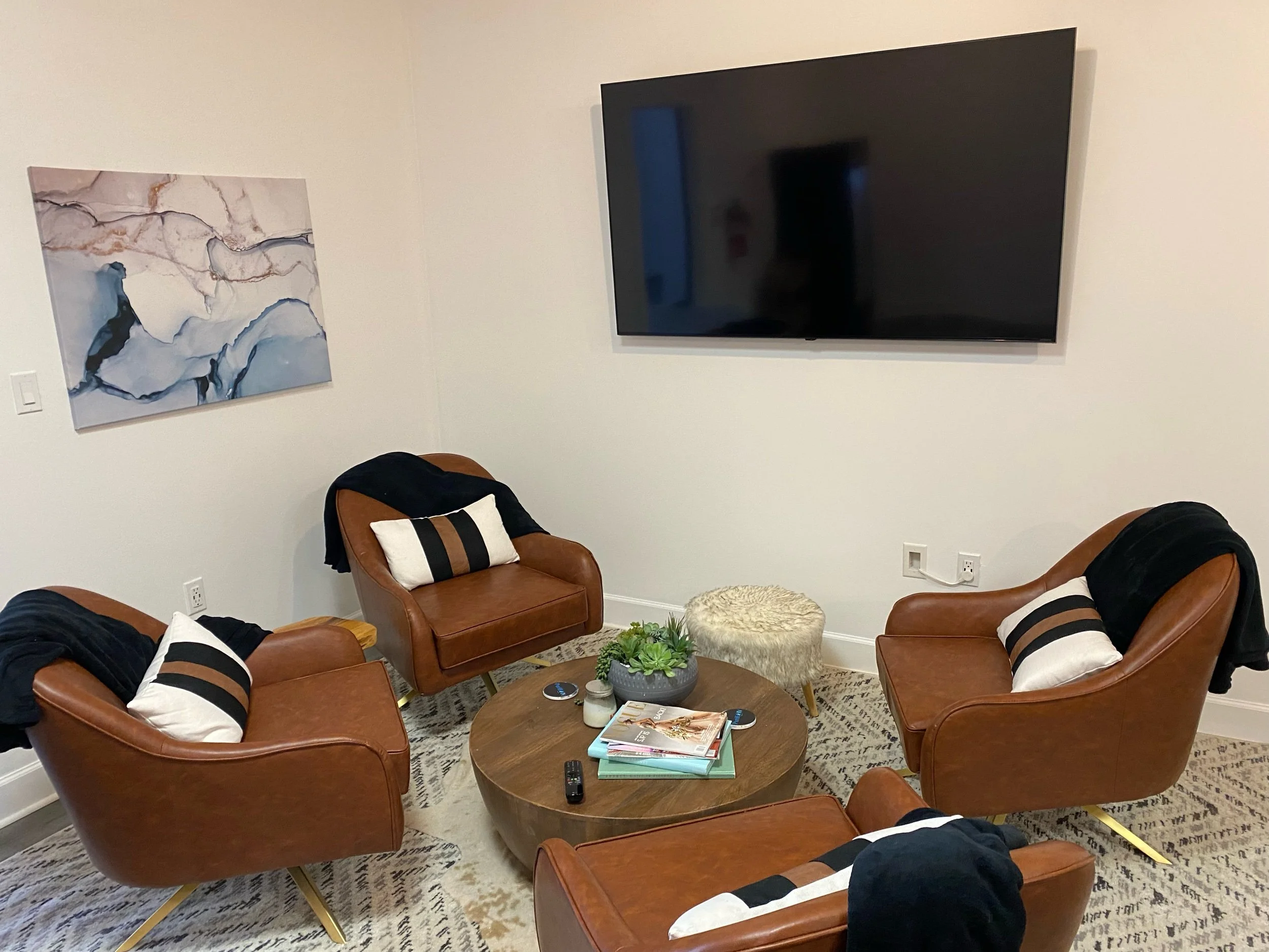 Cozy seating area with four brown leather chairs, a round coffee table, and a large wall-mounted TV. A decorative abstract painting is on the wall. Black and white cushions and throws add accent to the chairs. A plant and magazines are on the coffee
