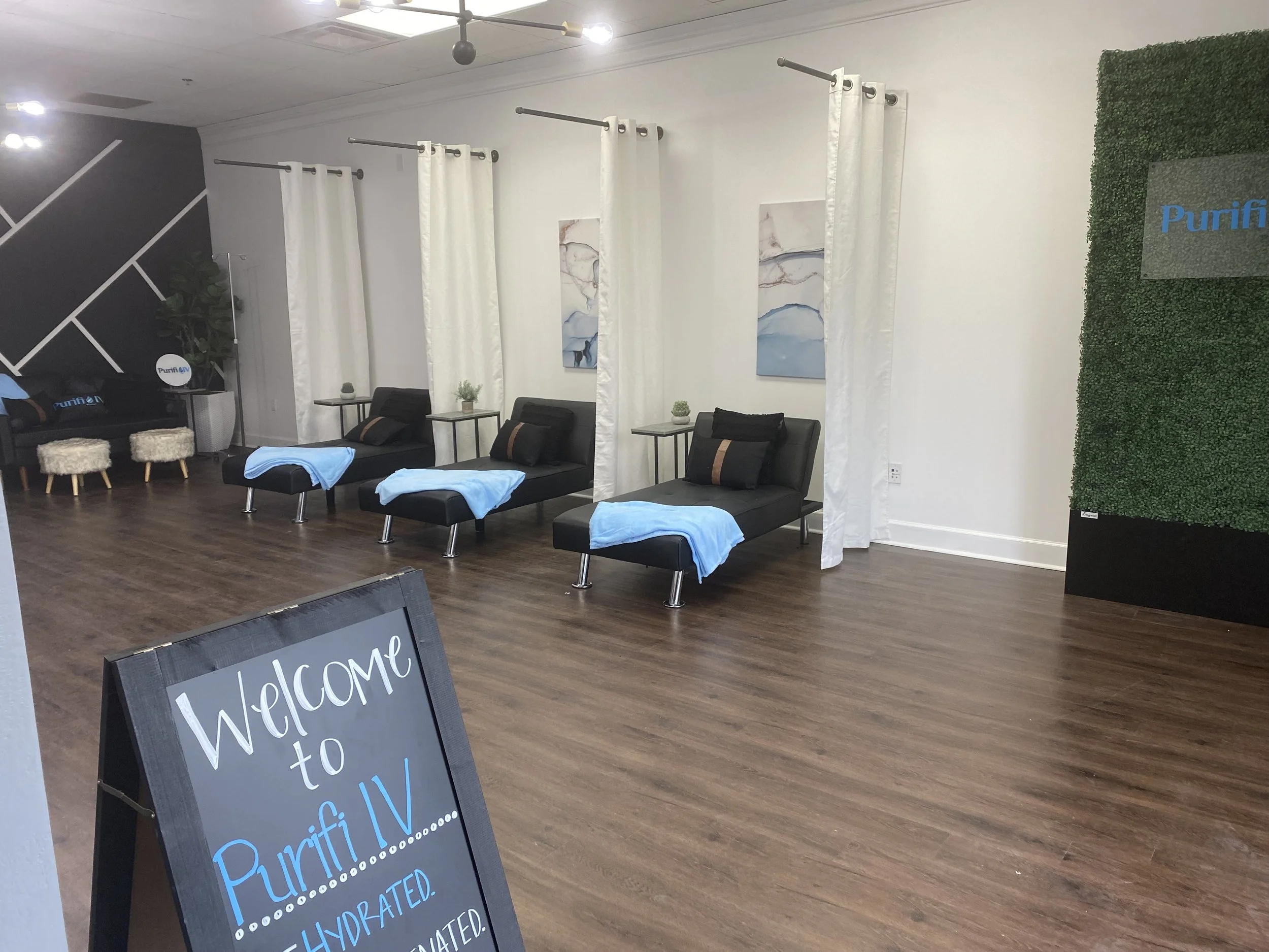 Interior of a wellness clinic featuring modern lounge chairs with light blue blankets, white curtains, a green wall with a sign, and a blackboard sign that reads 'Welcome to Purifi IV' with phrases 'hydrated' and 'elevated.' Wooden flooring and minim