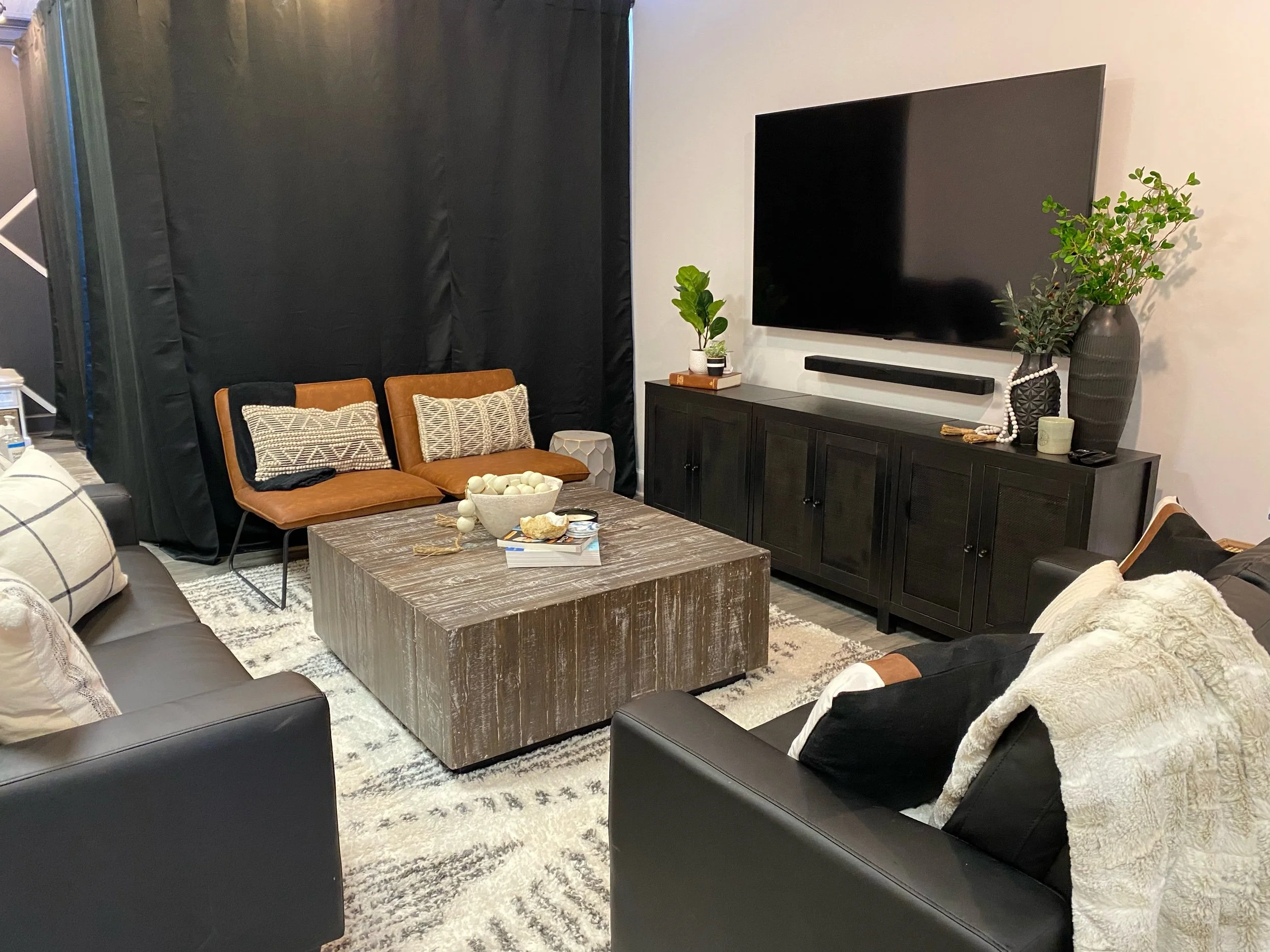 Cozy living room with modern decor, featuring a large flat-screen TV on a dark wooden console, a square wooden coffee table, leather chairs with patterned cushions, a black sofa, various plants, and decorative items.