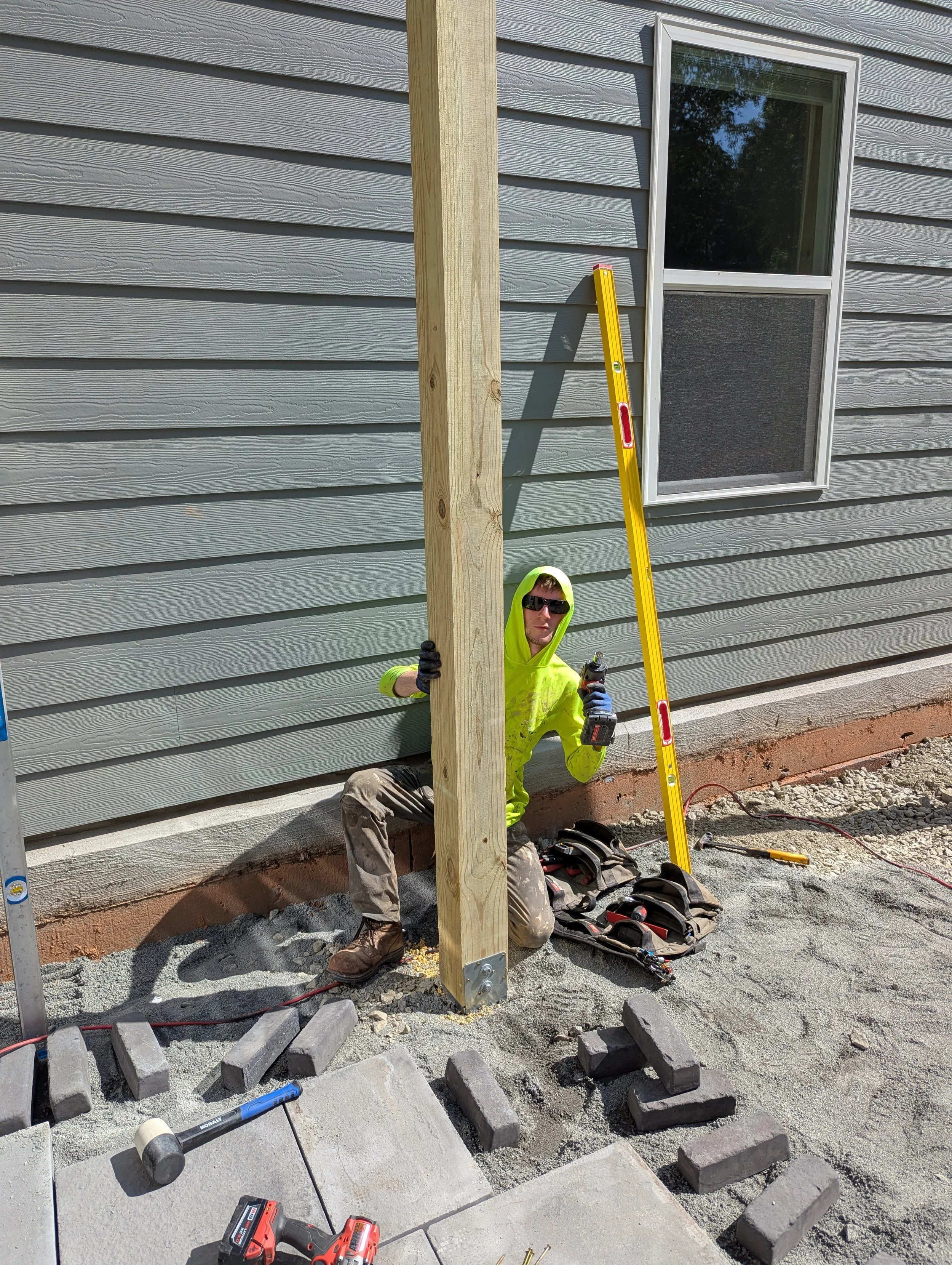A construction worker kneeling on the ground next to a wooden post, holding a power drill, with a level tool leaning against the house wall, near a window. The worker is wearing a bright yellow hoodie, sunglasses, gloves, and construction boots, surrounded by tools and bricks.