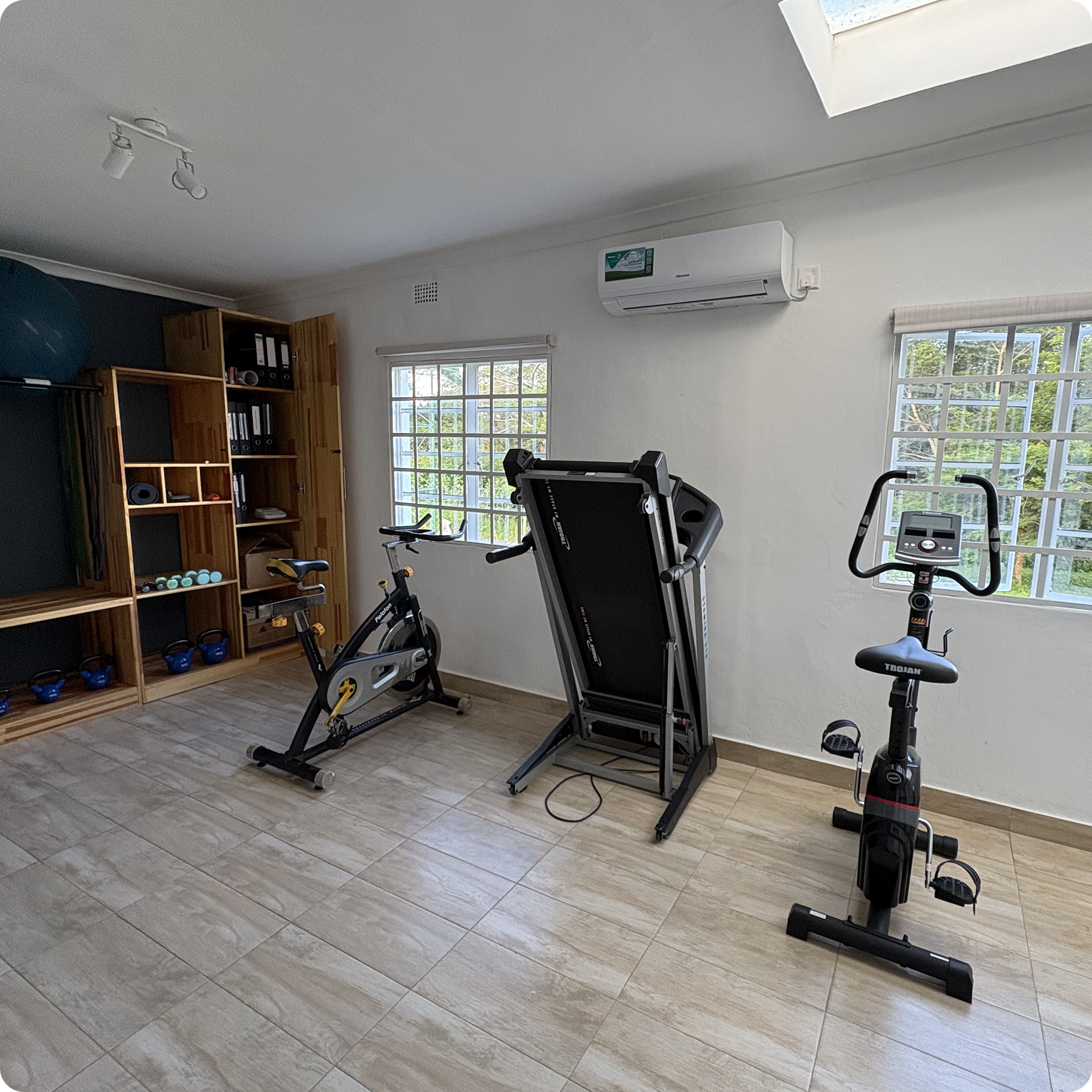Movement Therapy gym with exercise equipment including a stationary bike, a treadmill, and a compact exercise bike. There is a lot of natural light, a bookshelf with binders and kettlebells, and an air conditioning unit on the wall.
