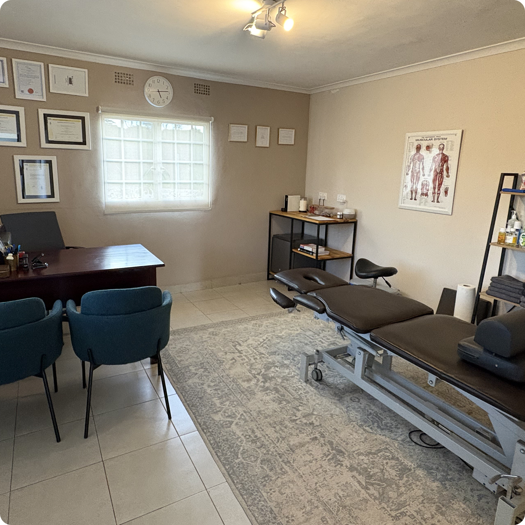 Movement Therapy examination and therapy room with a treatment table, a desk with a few chairs, framed certificates on the wall, a window, an air conditioner, and shelves with physiotherapy and medical supplies.