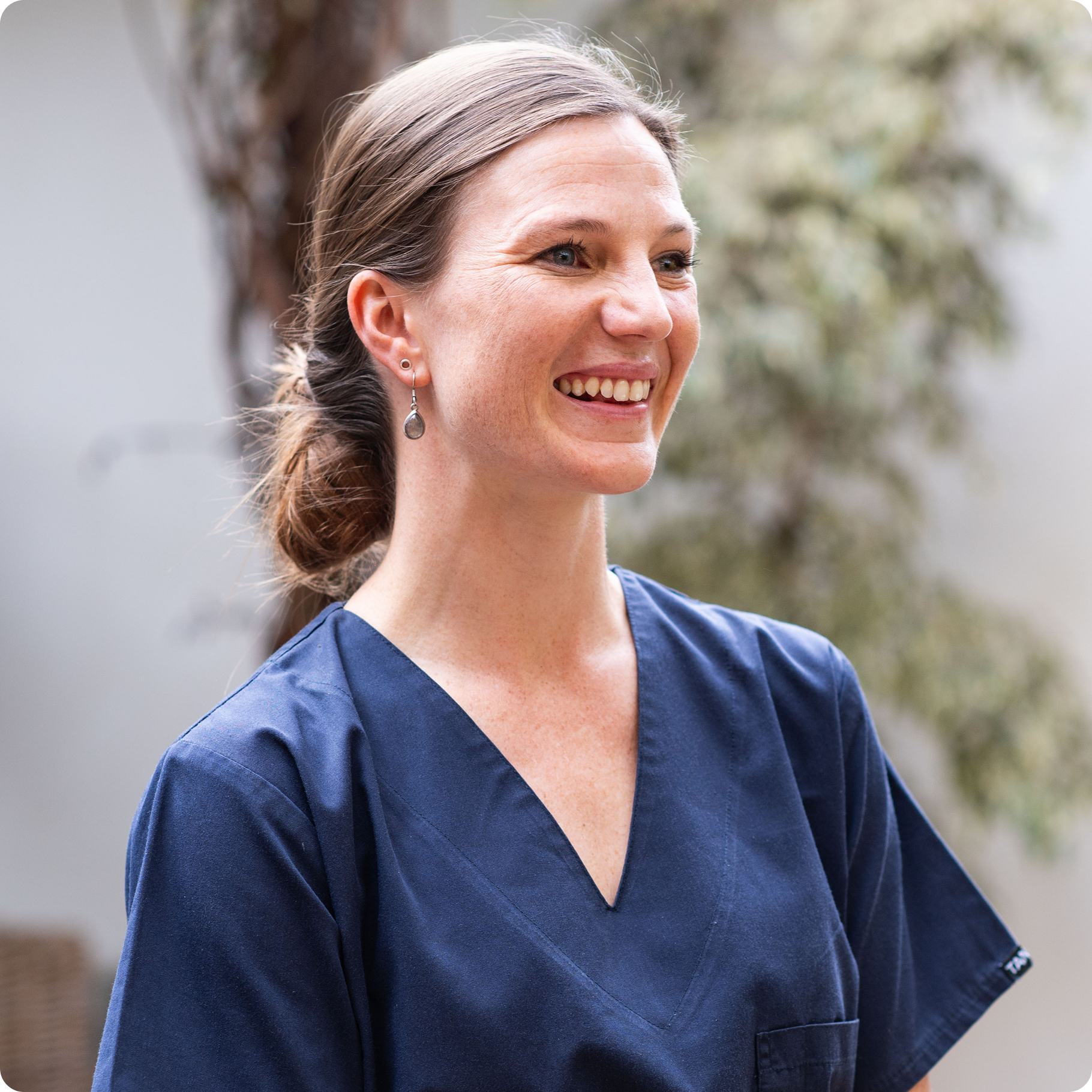 Michele Thomas, Specialist Physiotherapist shown smiling, wearing navy scrubs, with her hair tied back, standing outdoors with trees and a house in the background.