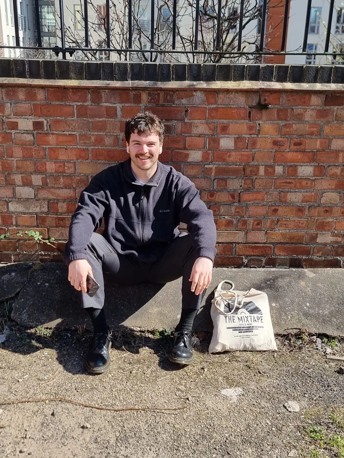 A young man with dark hair and a beard sitting on a concrete ledge in front of a brick wall, smiling at the camera. He is wearing a dark jacket, gray pants, and black shoes, holding a smartphone in his right hand. Next to him on the ground is a beige tote bag with the text 'THE MIXTAPE' and a headphone graphic.