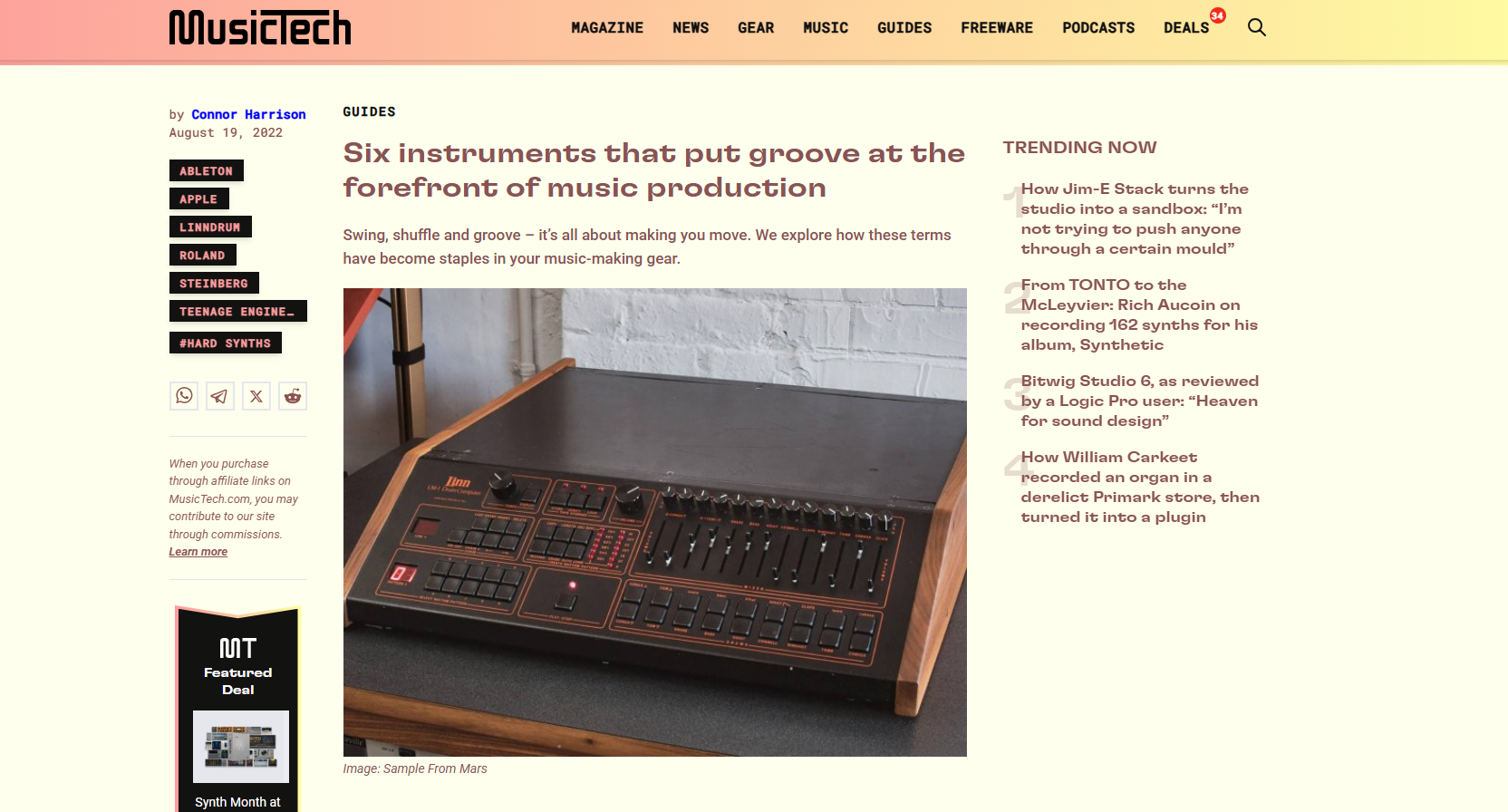 Photo of a vintage analog synthesizer or drum machine placed on a wooden table with a black surface, featuring knobs, sliders, and buttons. The background is a white brick wall.