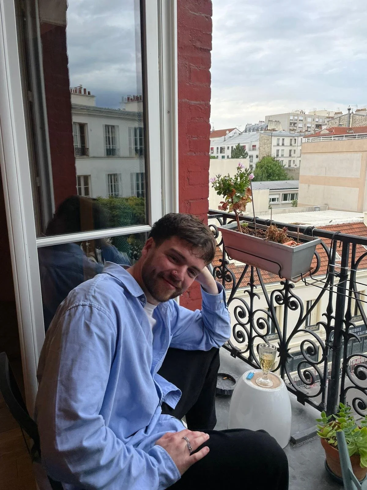 A man sitting on a balcony with a glass of sparkling drink on a small table. The balcony has decorative black iron railing, potted plants, and a city view in the background with cloudy skies.