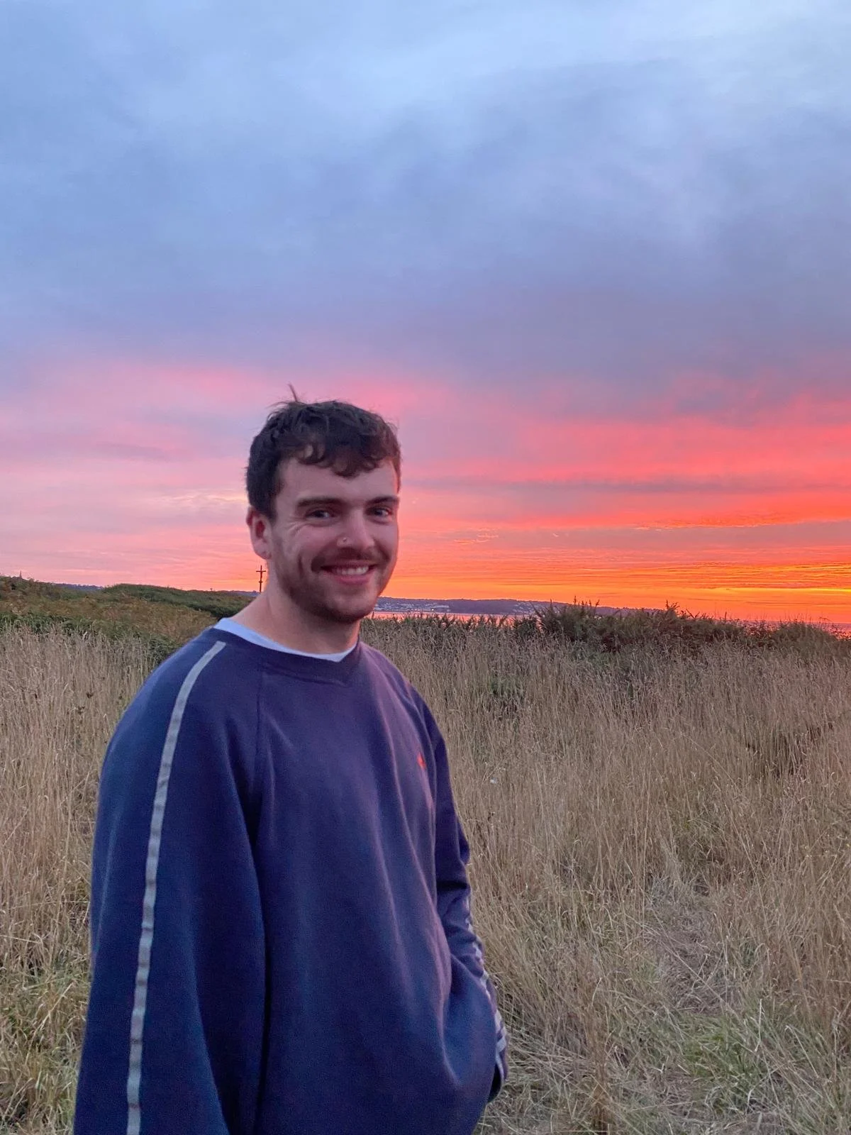 A young man smiling outdoors during sunset, standing in a grassy field with a colorful sky in the background.