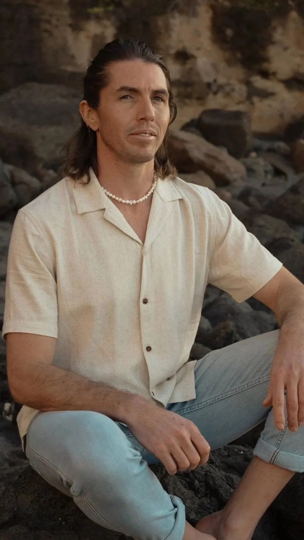 A young man with long brown hair, a nose piercing, and light skin is sitting on rocks at the beach during sunset. He is wearing a cream-colored short-sleeve button-up shirt, light blue rolled-up jeans. He is an Osteopath