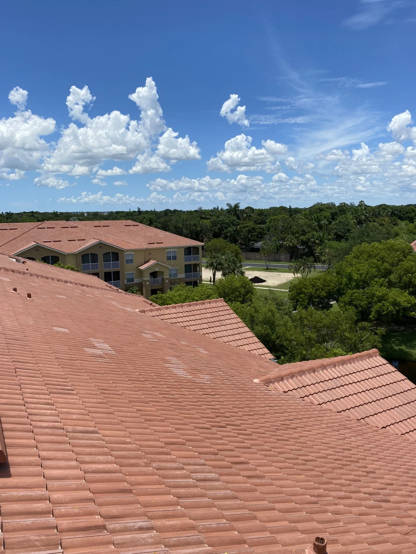 View of reddish clay tiled rooftops, with a background of trees, a building complex, and a blue sky with scattered white clouds.