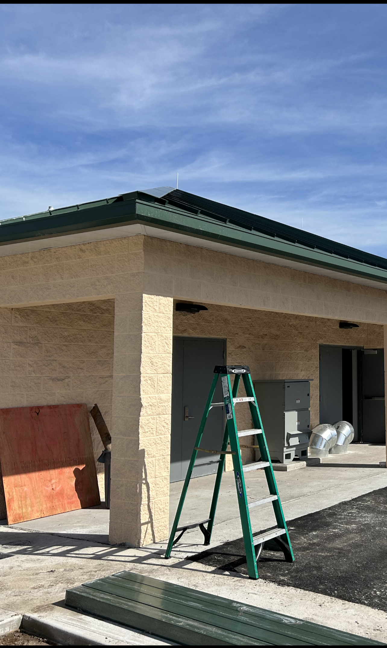 Exterior of a building with a green roof, beige brick walls, a green ladder, construction materials, and a blue sky with clouds.