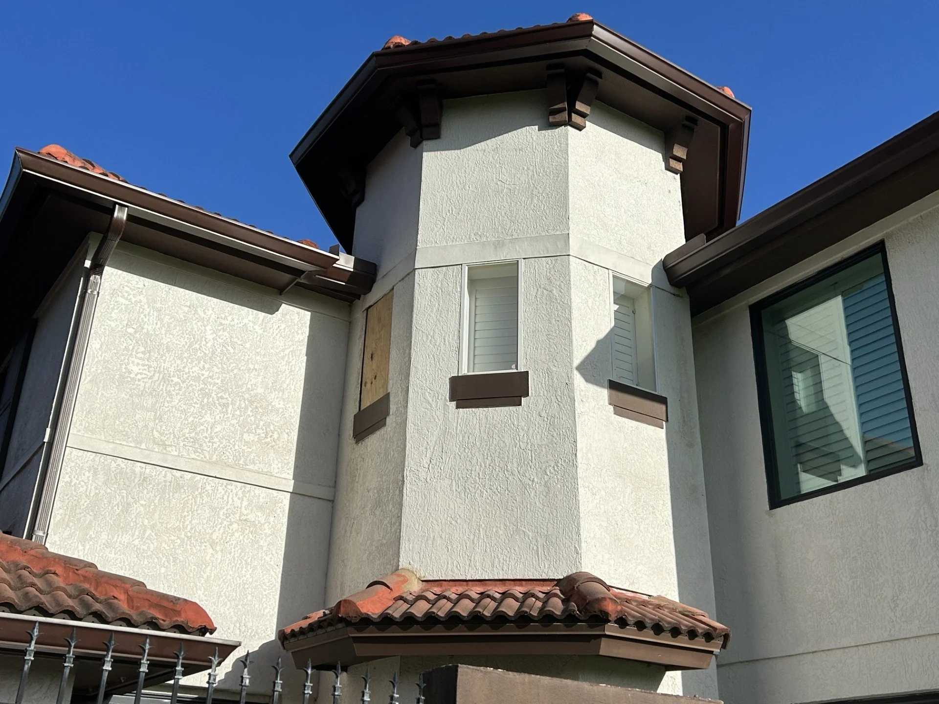 Close-up of a house's exterior showing a white stucco wall with a small tower-like structure, brown trim, and a red tile roof under a clear blue sky.