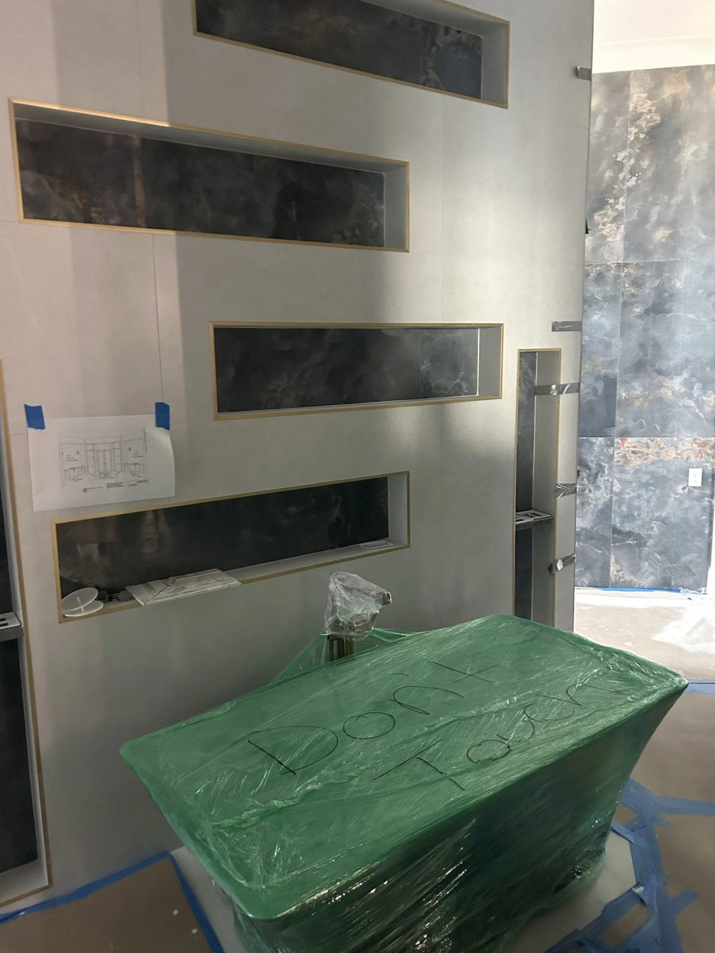Interior view of a construction site with a large green wrapped object labeled in black marker, tall rectangular wall niches with black marble inserts, and a textured stone wall in the background.