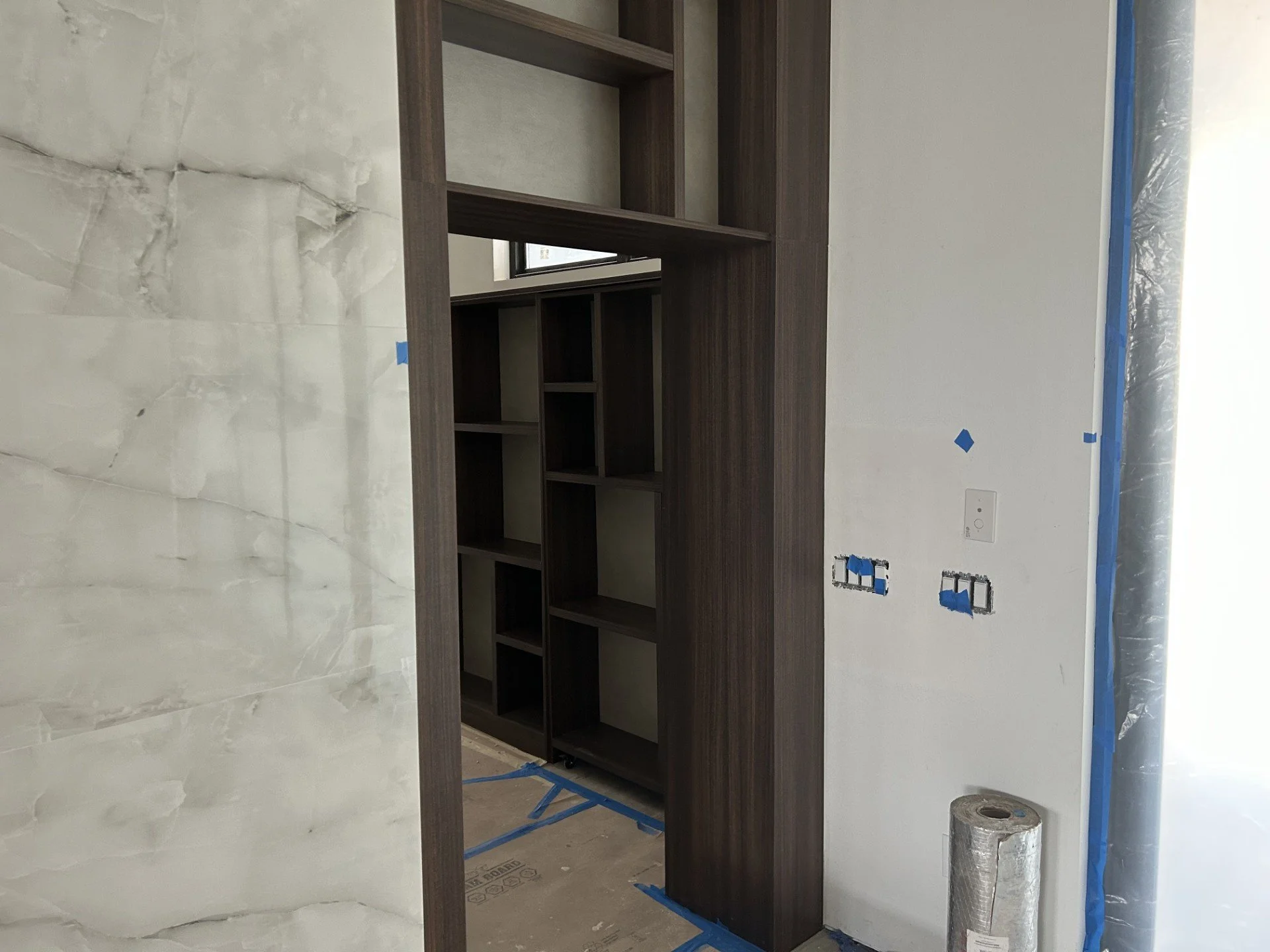 Interior view of a room under construction showing a wooden shelving unit and unfinished wall with electrical outlets and coverings.