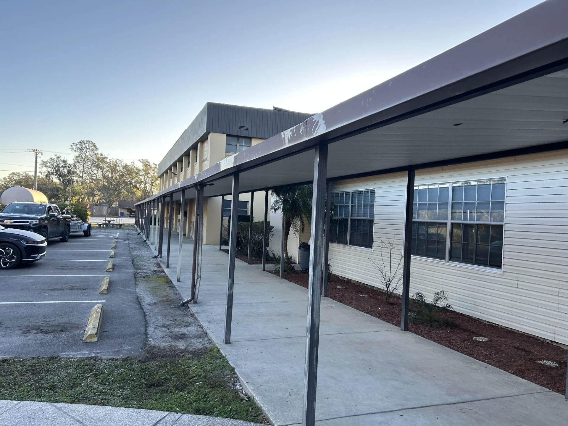 Side view of a commercial building with a covered walkway supported by metal posts, windows, and parked cars in a lot.