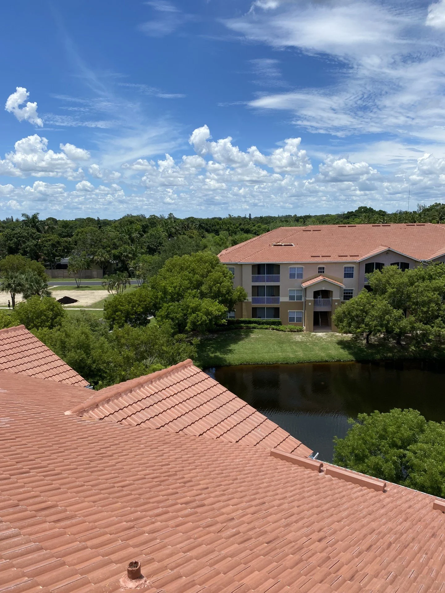 View of rooftops with red tiles, trees, a small pond, and a multi-story apartment building in the background under a partly cloudy sky.