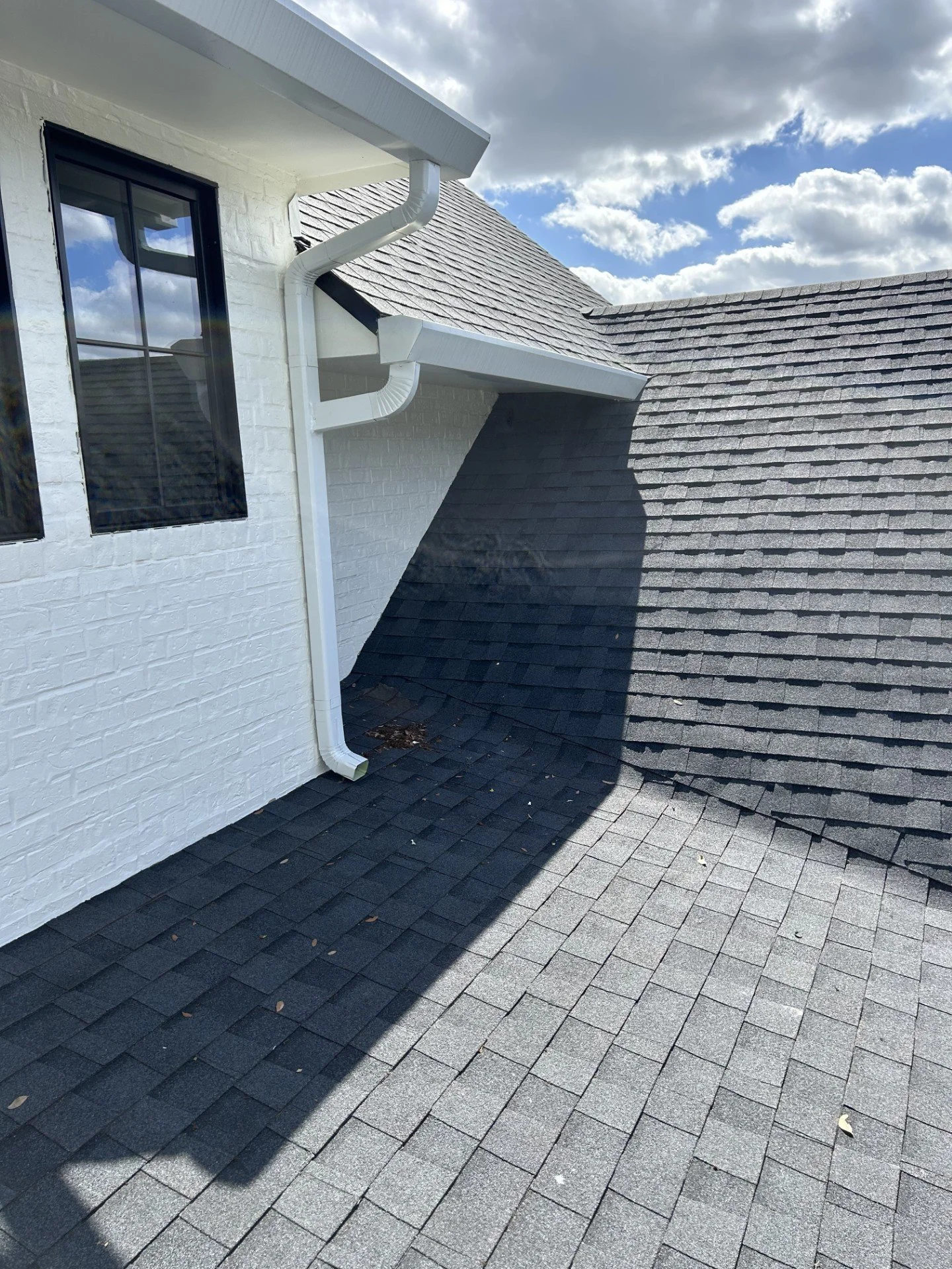Close-up of a roof with two different shades of shingles, a white brick wall with two windows, and white gutters, under a sky with clouds.
