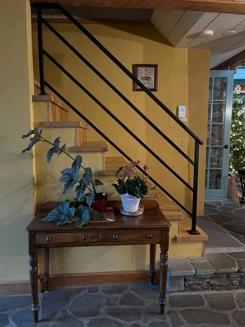 Interior view of a stairway landing with a decorative black metal railing, a window with a chandelier hanging above, and some construction tape on the floor indicating ongoing work.