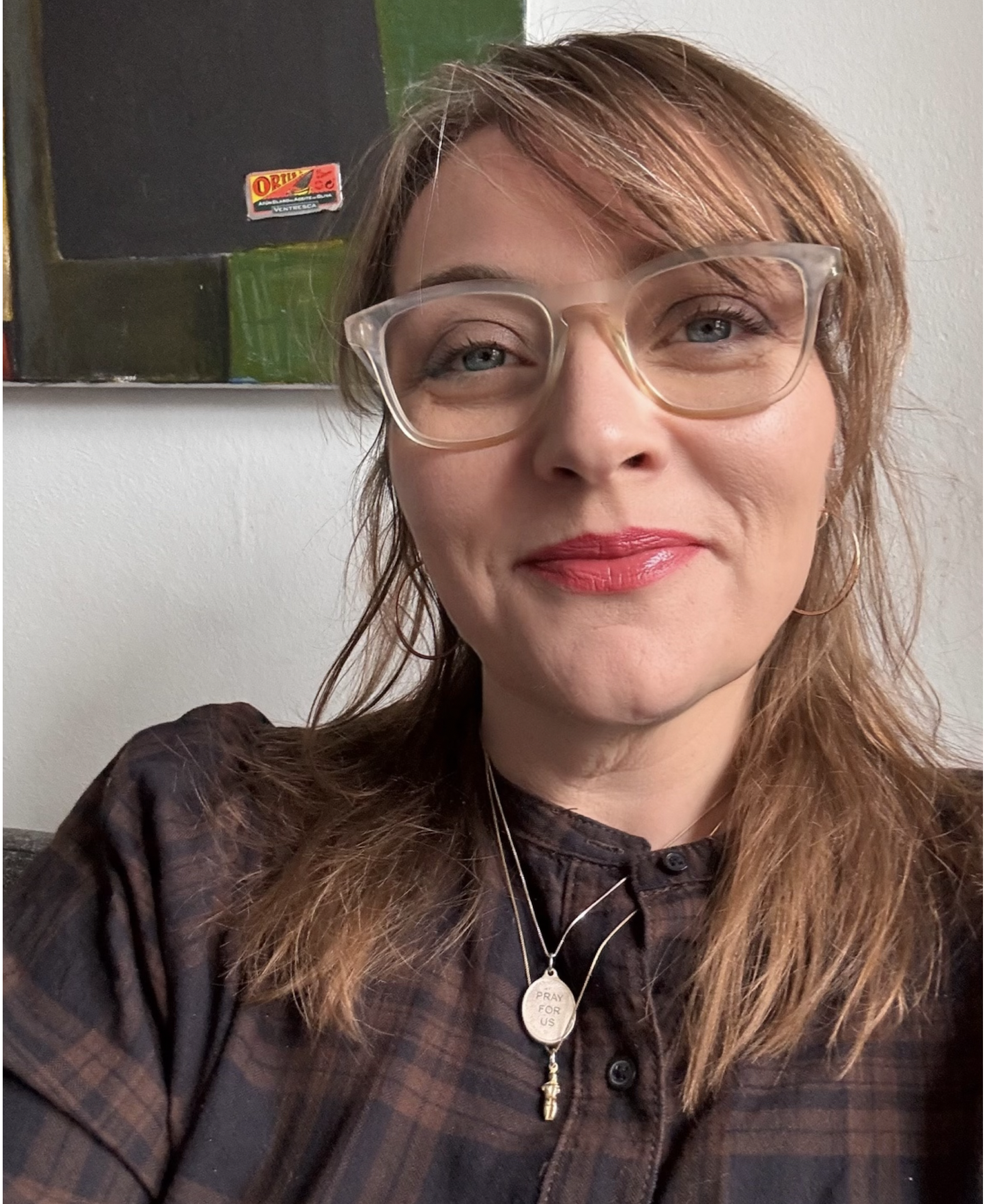 A photo of Claire Doherty. She has light brown hair, is wearing large, clear glasses, pink lipstick and hoop earrings, smiling slightly in front of a white wall and a blackboard.
