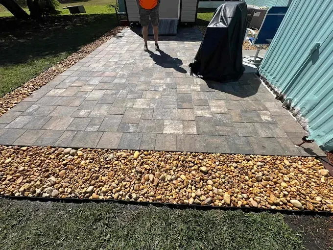 Paved backyard area with a gravel border, a person standing, and outdoor furniture, next to a light blue metal shed.