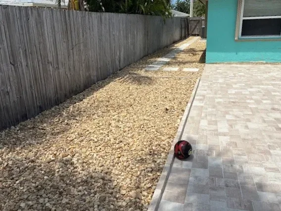 Backyard with a gravel path, a wooden fence, a turquoise house wall, and a red and black ball on the paved area.