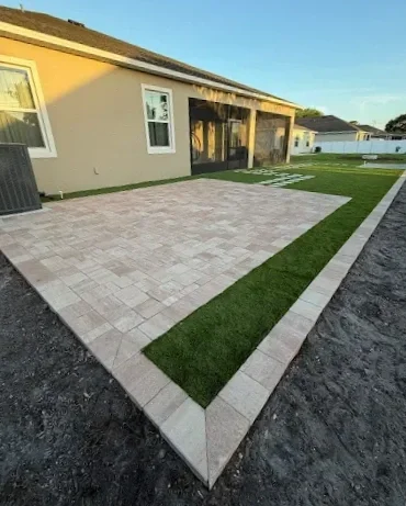 Backyard with a paved patio, bordered by green grass, in front of a beige house with white-framed windows.