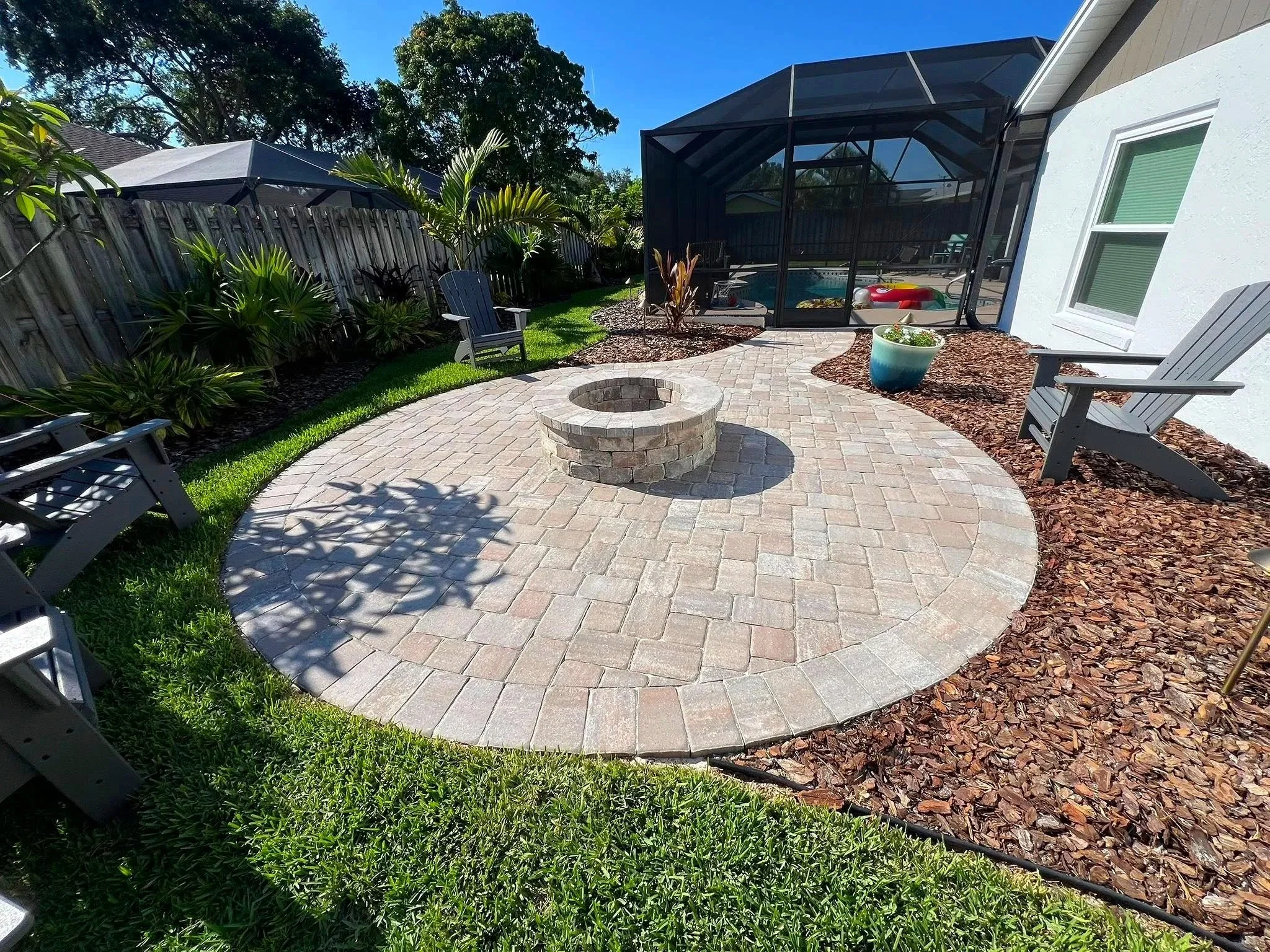 Backyard patio with brick pavers, green grass, garden beds, and plantings, enclosed by a wooden fence. There are Adirondack chairs, a fire pit, and a screened-in pool area with a covered structure. The sky is clear and sunny.
