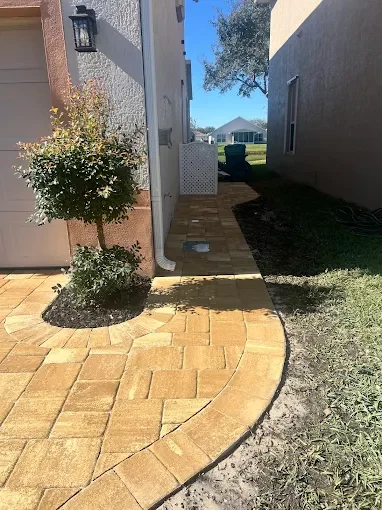 Backyard path with brick pavers, small tree, white lattice fence, and neighboring house in the distance.