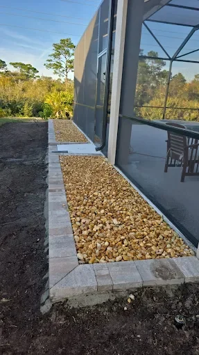 A row of large rectangular pavers framing a gravel bed alongside a building with large windows, with trees and blue sky in the background.