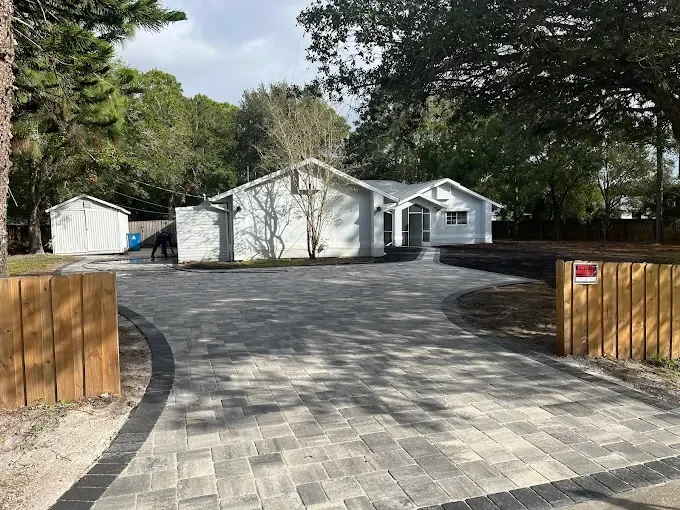 Newly paved driveway leading to a white house with trees in the background and wooden fencing on both sides.