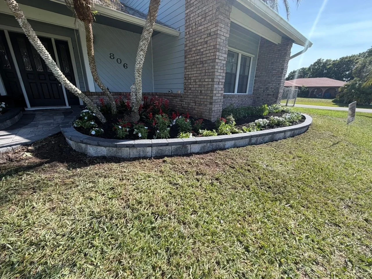 Front yard of a house with a flower bed bordered by grey bricks, containing a small tree, white and red flowers, and green bushes, with a grassy lawn in the foreground and neighboring houses in the background.