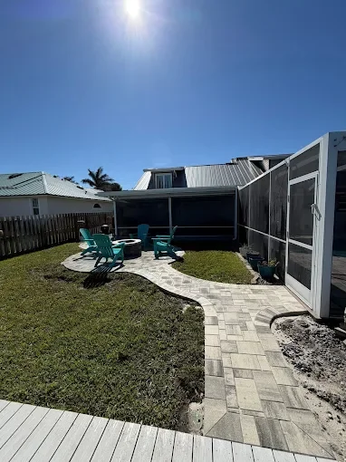 Backyard with a paved pathway, a grassy area, four turquoise chairs around a small table, a screened porch, and neighboring houses under a clear blue sky.