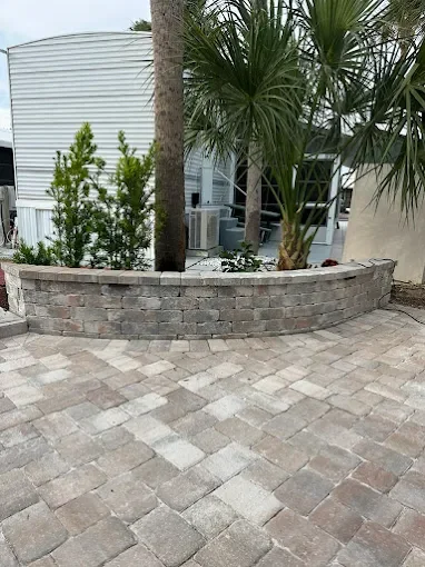 A landscaped backyard area with a curved brick retaining wall, palm trees, other plants, and a paved stone patio.