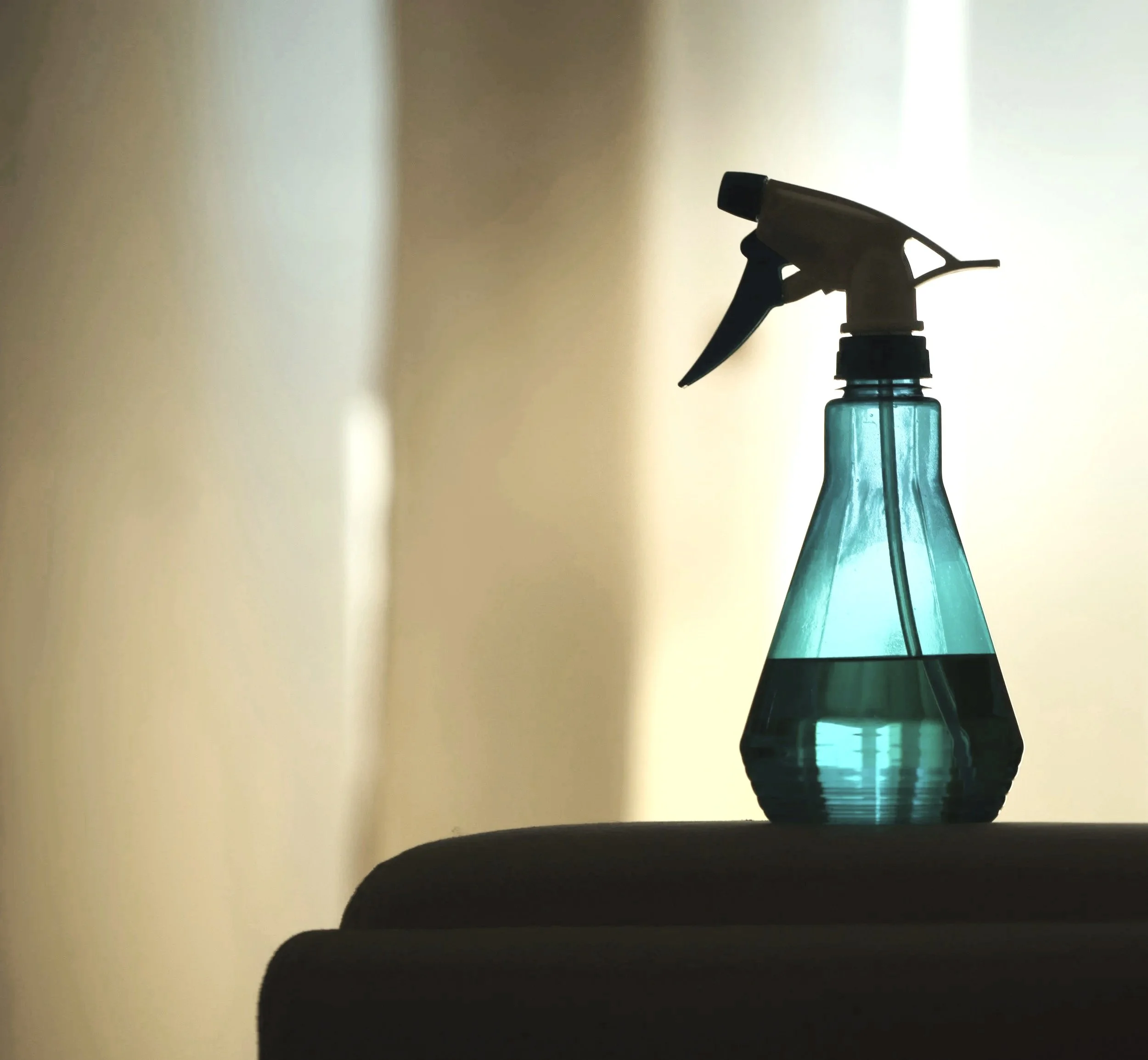 A spray bottle with a black trigger and a blue-green transparent body filled with liquid, placed on a dark surface, with a bright, softly lit background.